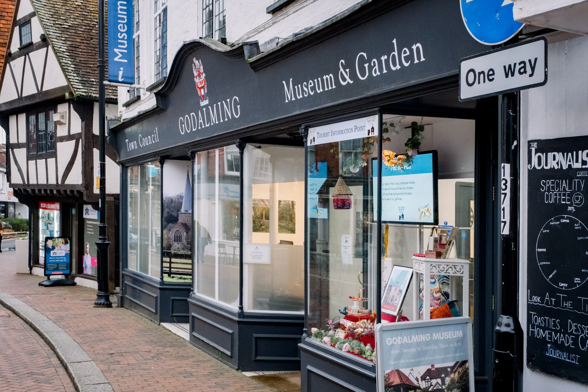 Street view of Godalming Museum and Garden with a black storefront, signage indicating it is a museum and tourist information point, and nearby buildings including a Tudor-style structure. A one-way sign is visible, along with display boards and a sidewalk with a brick pattern.