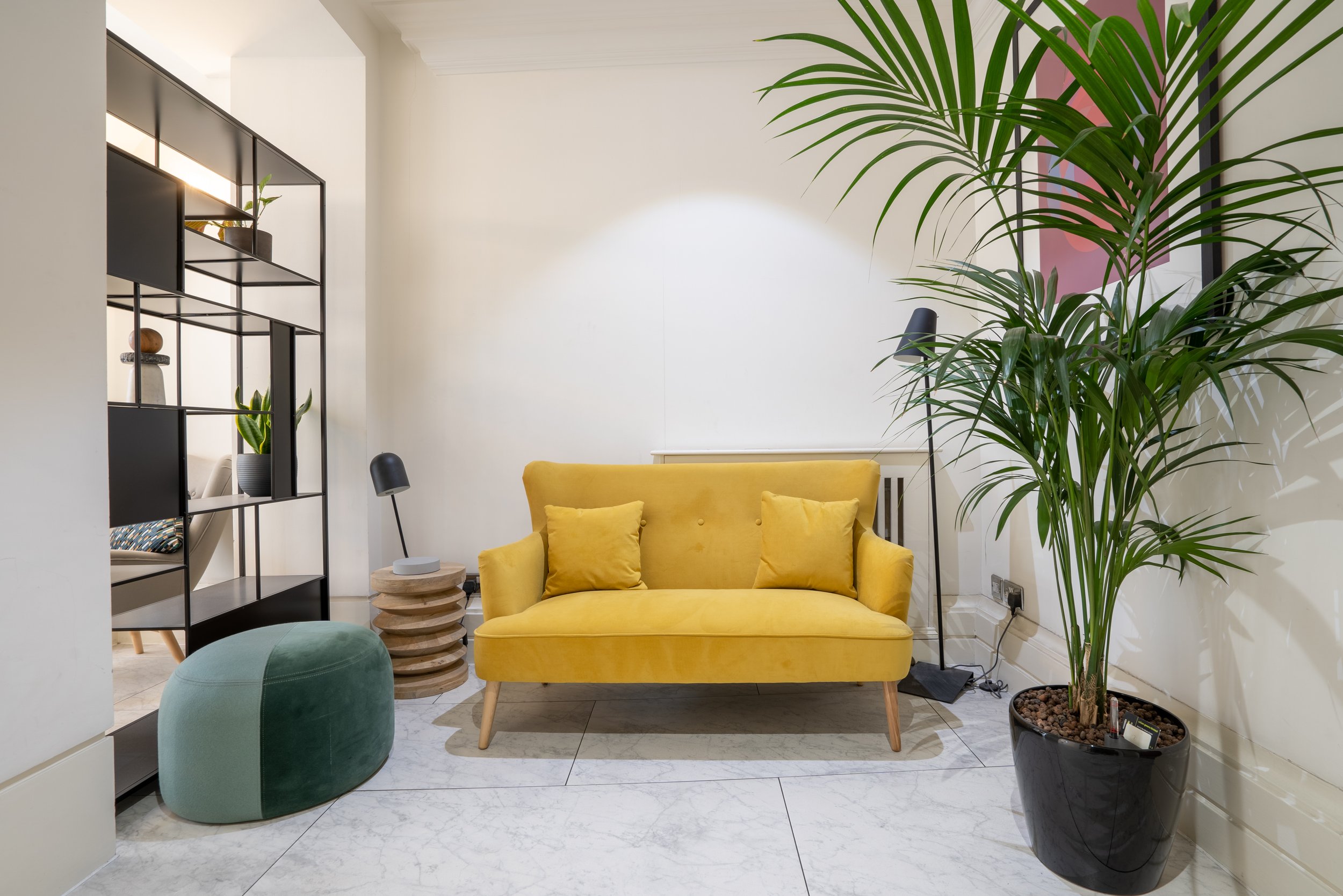 Modern living room with a yellow sofa, large potted palm, green ottoman, black shelving unit with decorative items, and a small side table with a black desk lamp, white walls, and marble flooring.