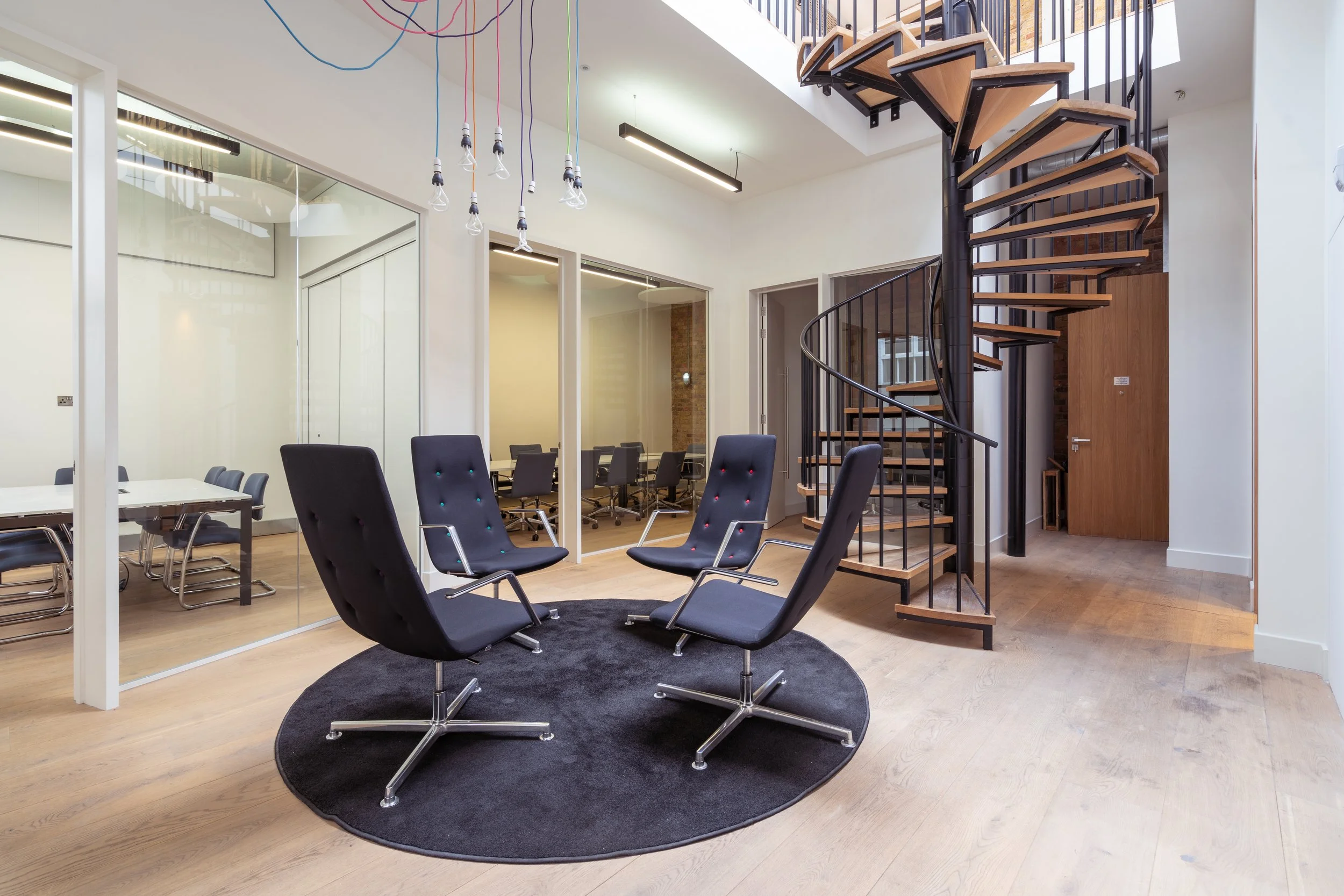 Modern office meeting room with four black chairs on a round black rug, a glass-walled conference room with additional chairs, and a wooden spiral staircase leading upstairs.