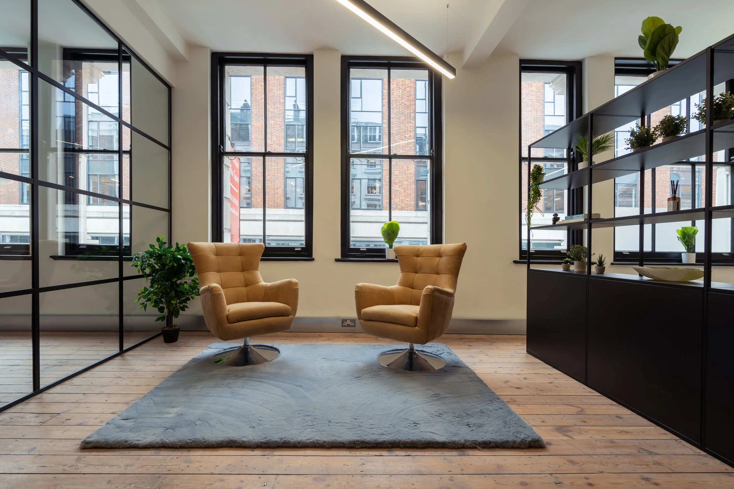 Modern office space with two beige armchairs, a gray rug, a large black shelving unit with plants, and three large windows showing brick buildings outside.