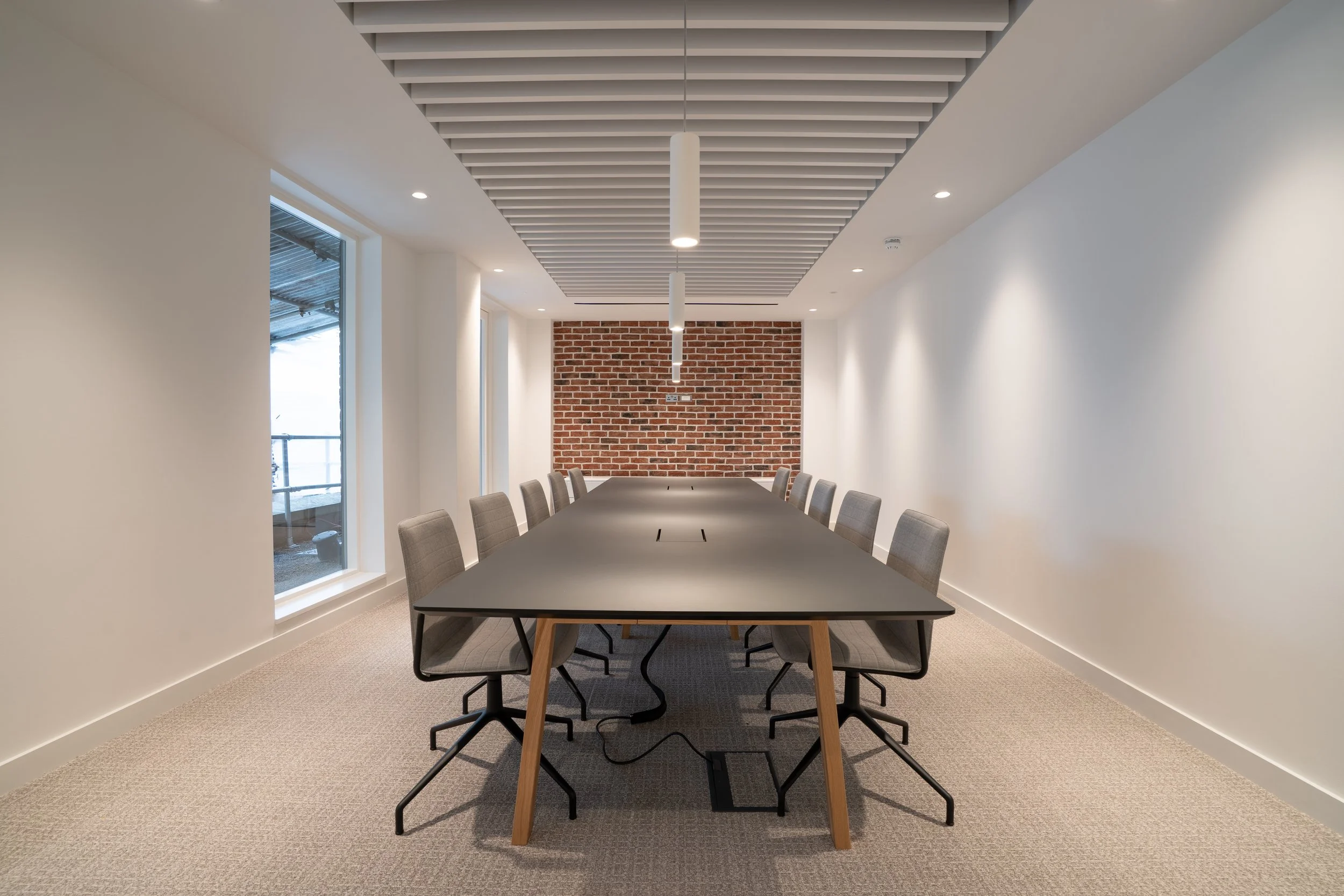 Modern conference room with a large black table, gray chairs, brick accent wall at the back, white walls, and ceiling with suspended beams and hanging lights.