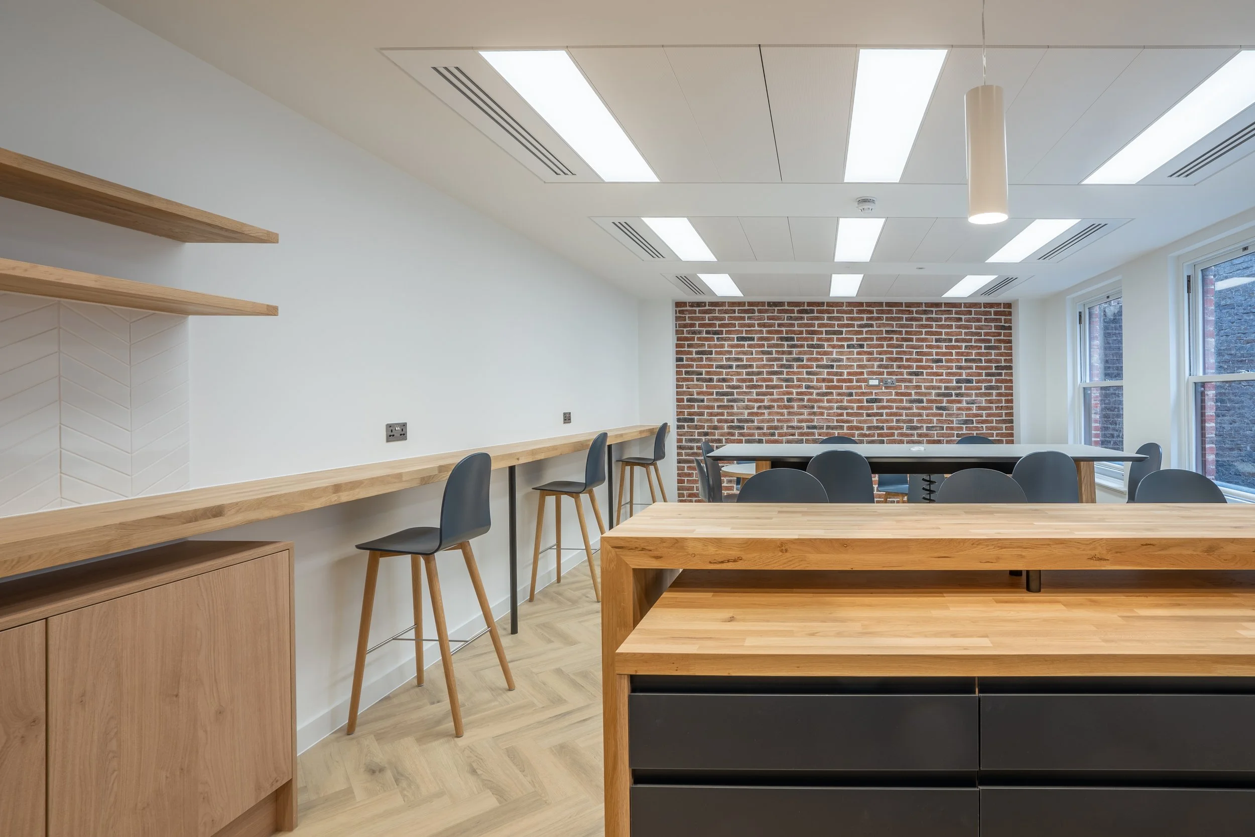 Modern interior room with wooden tables, chairs, brick and white walls, large windows, and ceiling lighting.