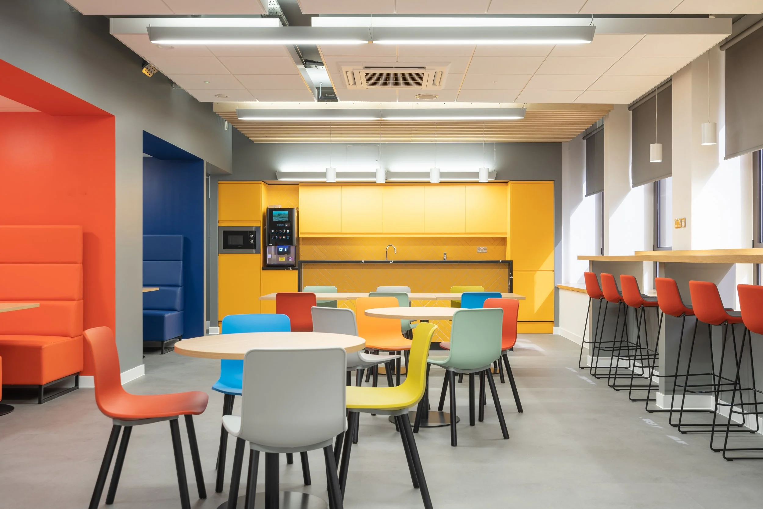 Colorful modern break room with tables, chairs, kitchen area with yellow cabinets, and large windows.