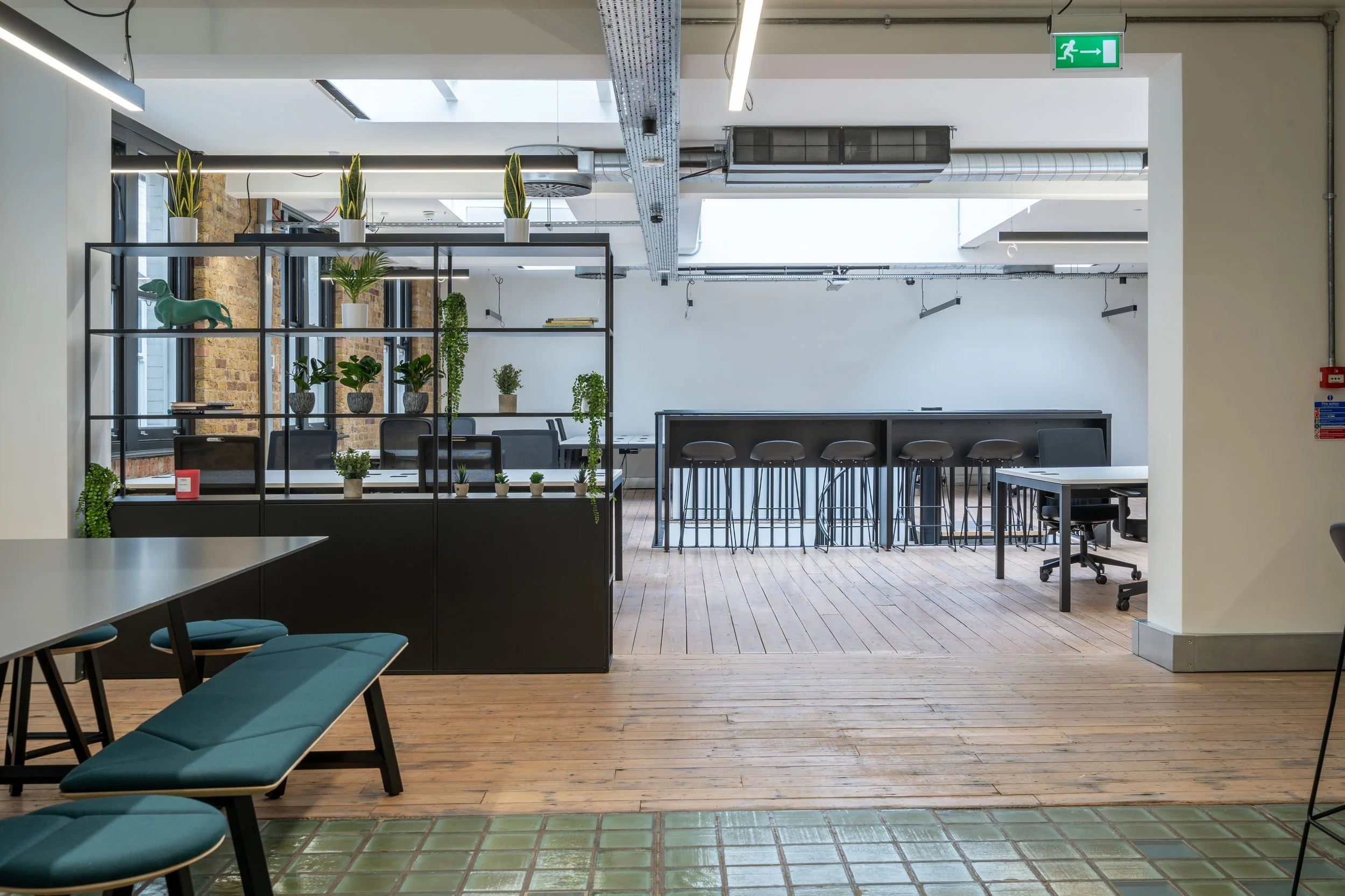 Interior of a modern office with wooden floors, black furniture, potted plants, and a glass wall section. An emergency exit sign is visible at the top right corner.