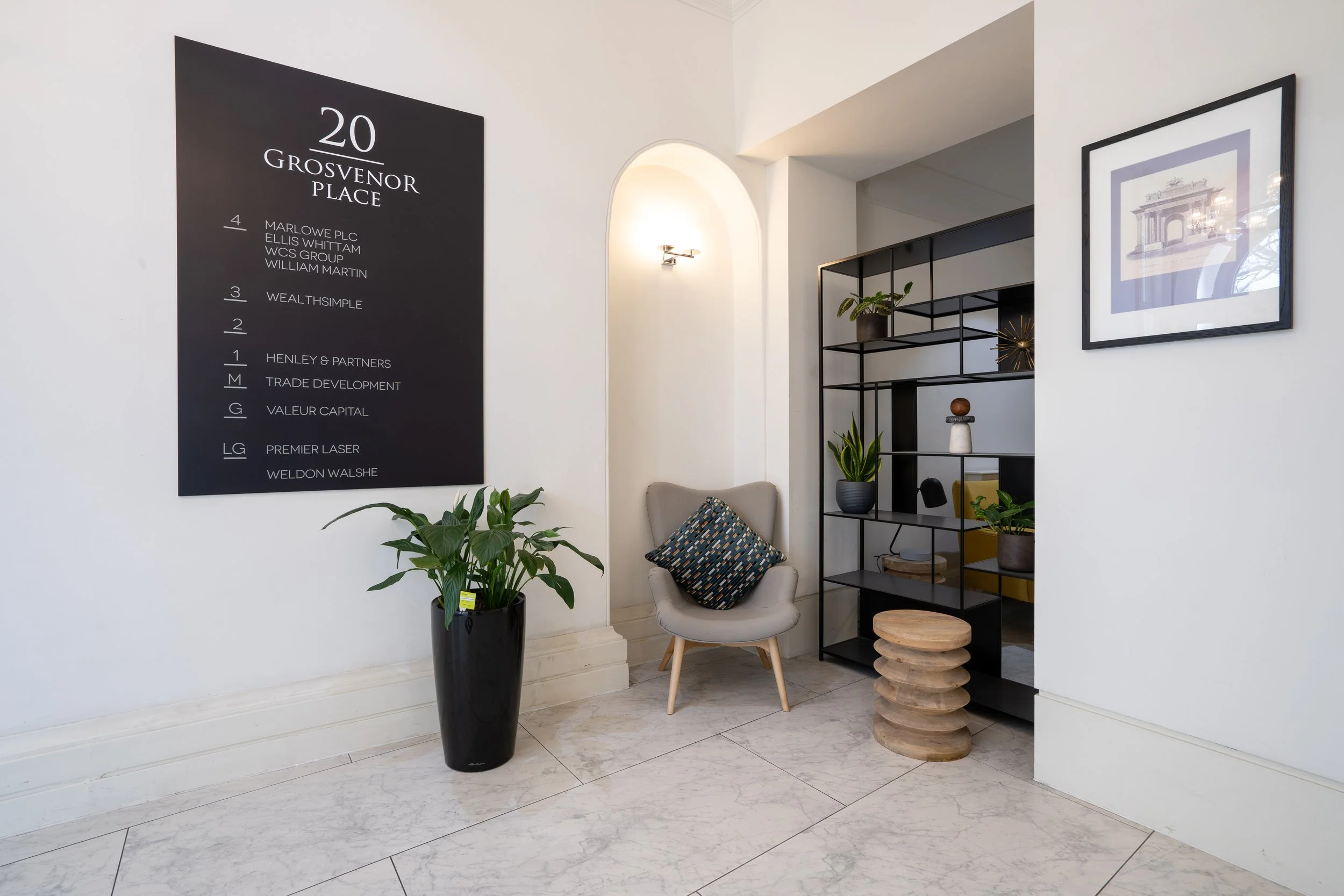 Lobby of a building with a black directory board, a potted plant, a modern chair with a cushion, and a black metal shelving unit with plants and decorative items, and a framed artwork on the wall.