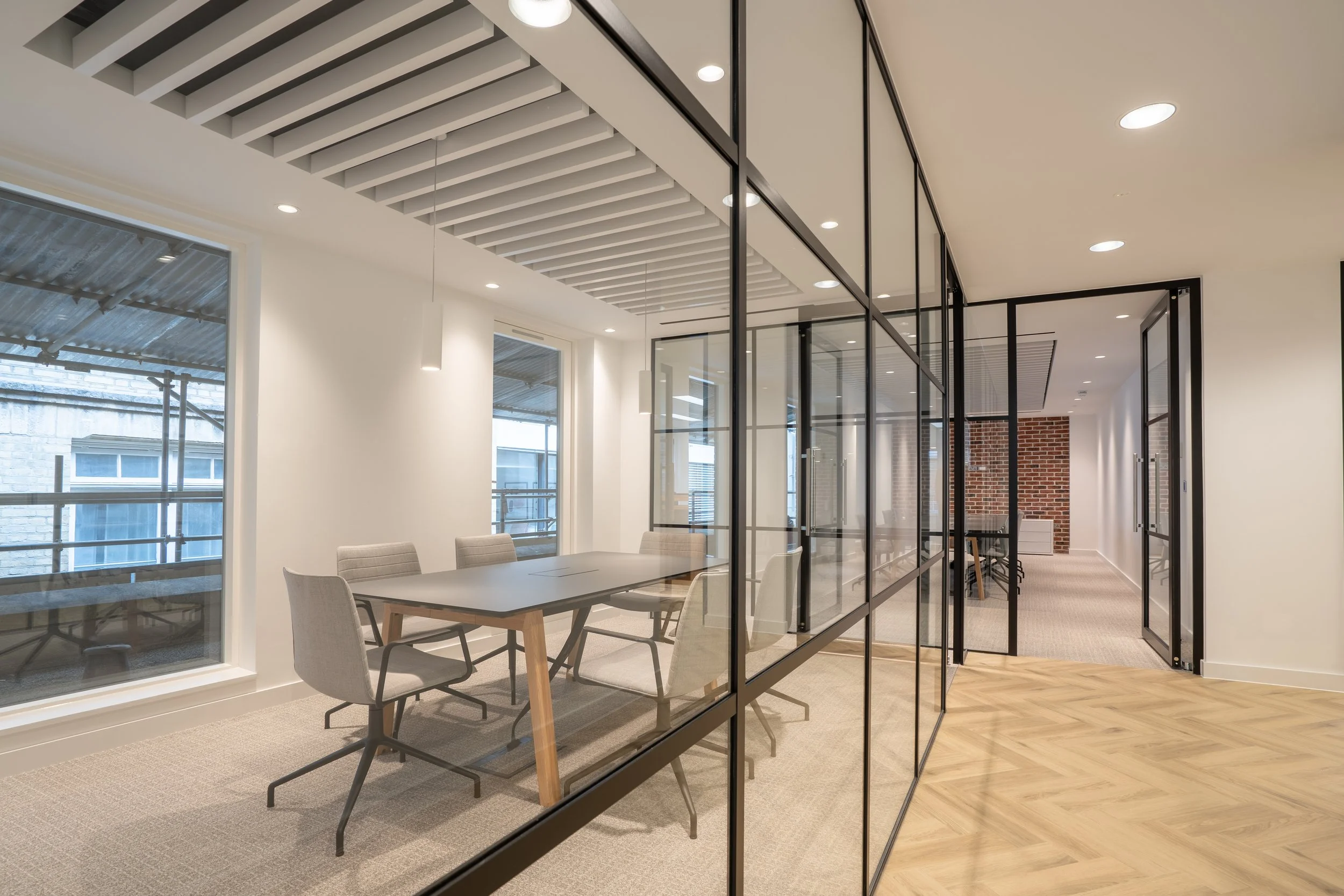 Modern office conference room with a glass wall, a grey table, six chairs, large windows, and ceiling lights.