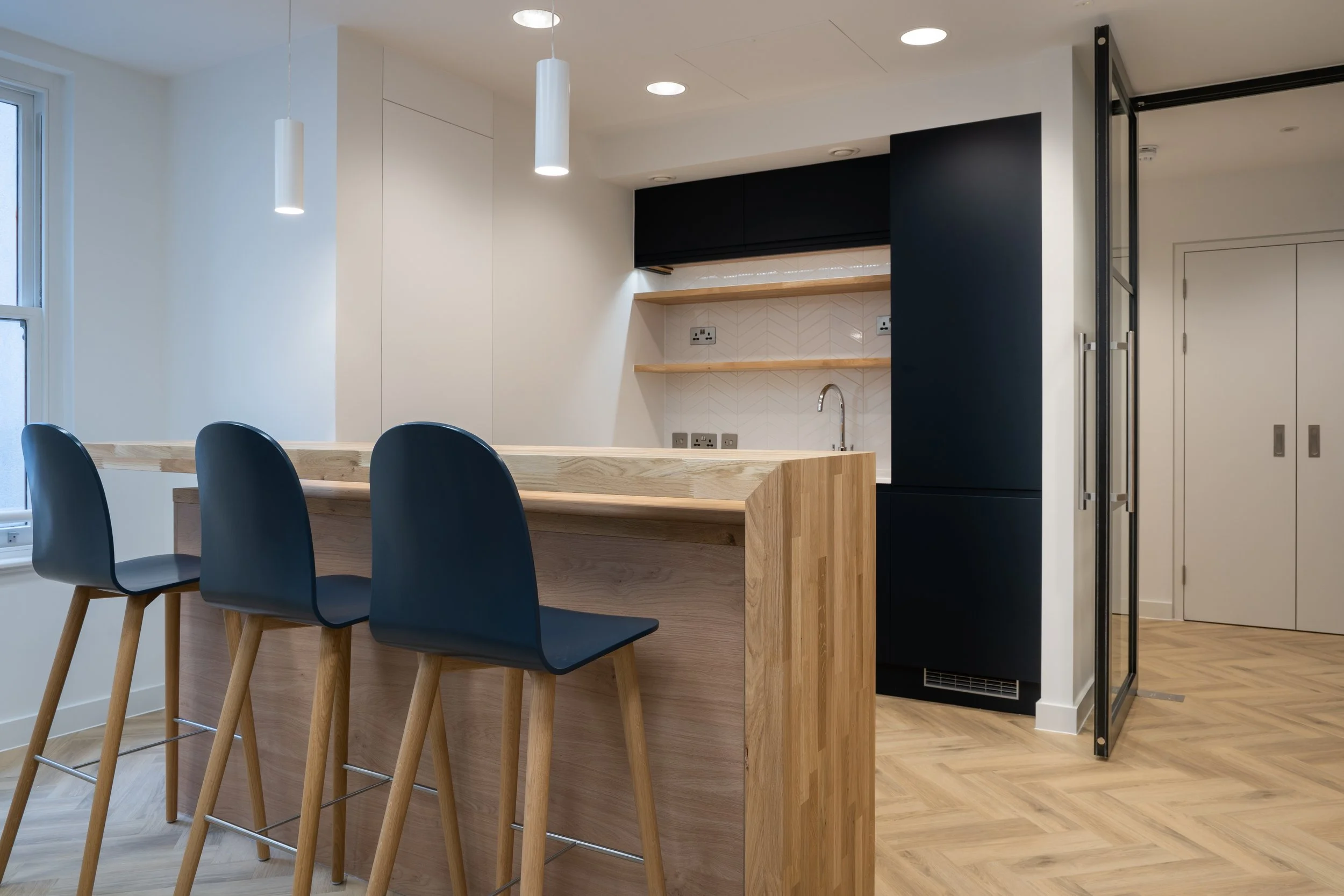 Modern kitchen with a wooden island and three black chairs with wooden legs, white walls, black cabinetry, open shelving, a sink, and a herringbone wooden floor.