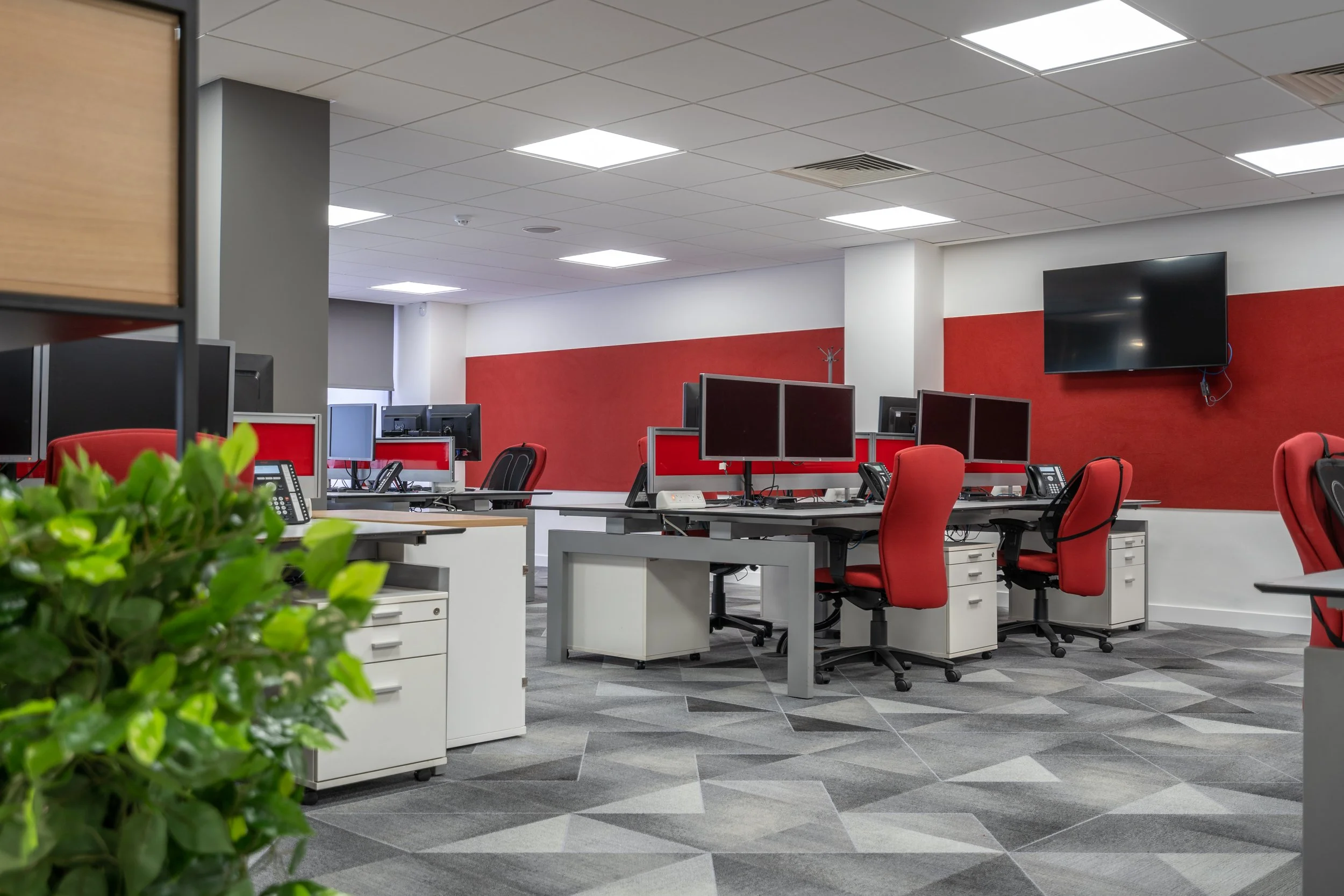 Empty modern office with red chairs, computer desks with multiple monitors, and a large wall-mounted TV, with a gray, red, and white color scheme.