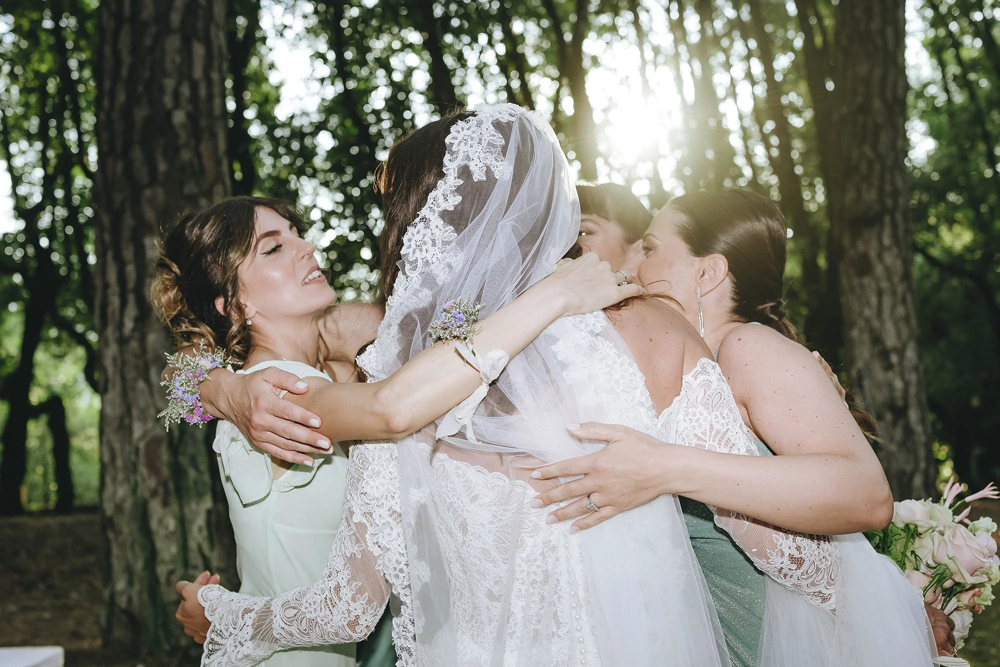 A bride hugging and being embraced by three women outdoors in a wooded area, sunlight filtering through the trees.