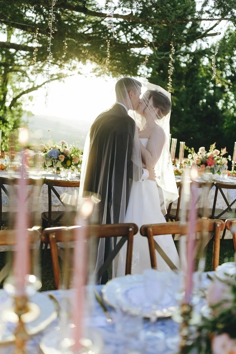 A bride and groom share a kiss at an outdoor wedding reception with decorated tables and a lush green background.
