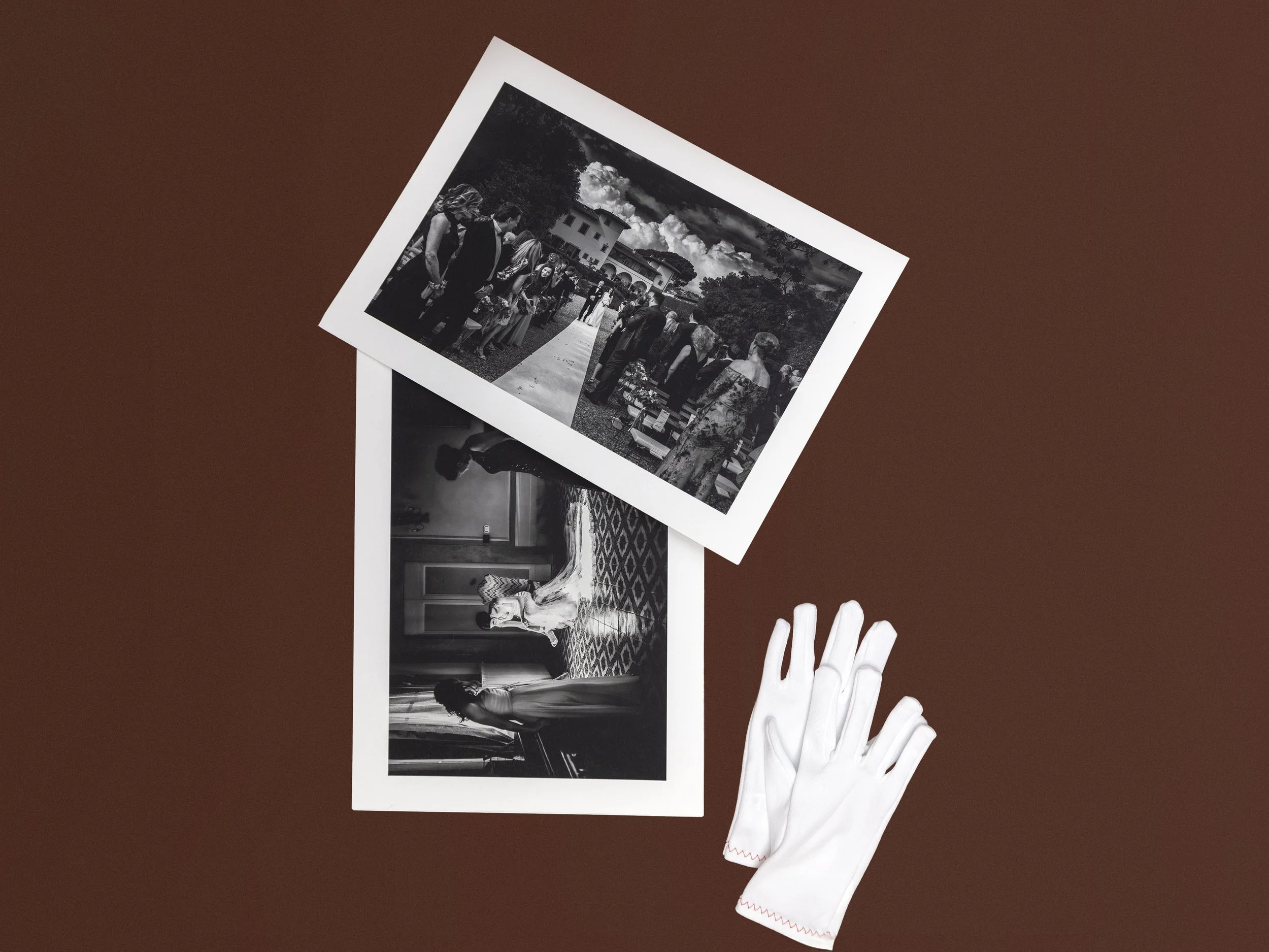 Two black and white photographs, one showing a ceremony with people on a sidewalk and the other showing women in formal dresses indoors, with a pair of white gloves nearby, all on a brown surface.