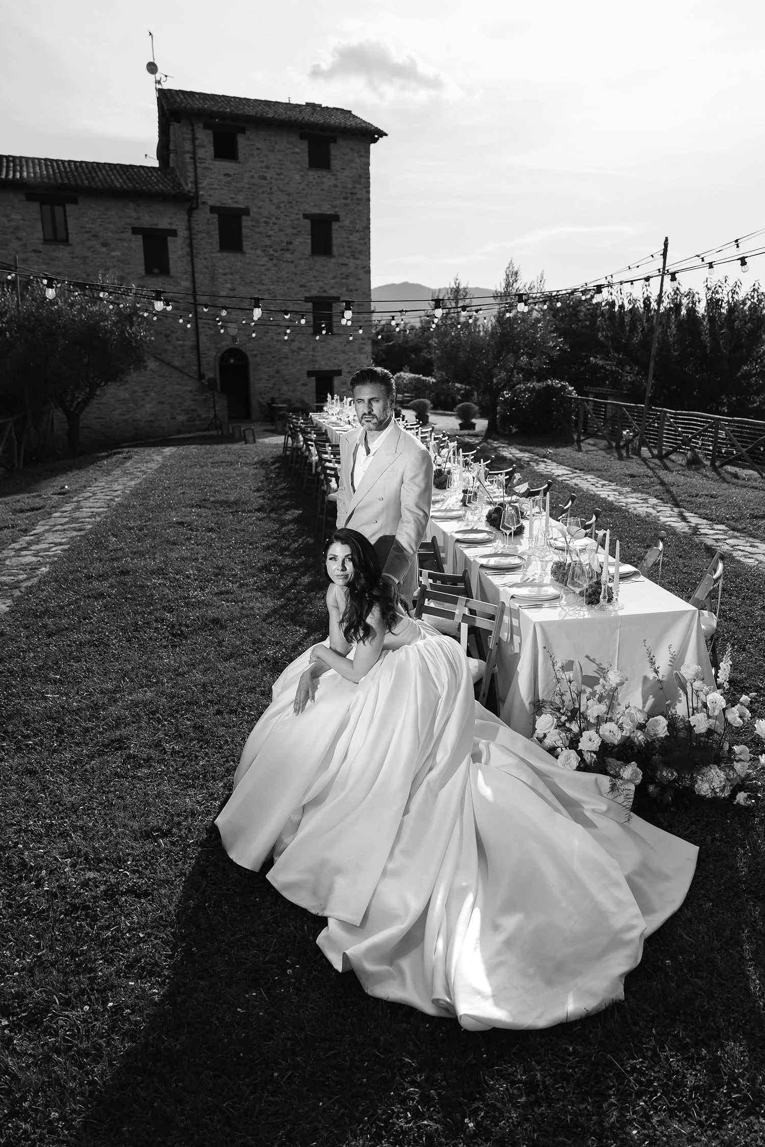 portrait of the bride sitting on a chair at the dinner tables with the groom standing in the back