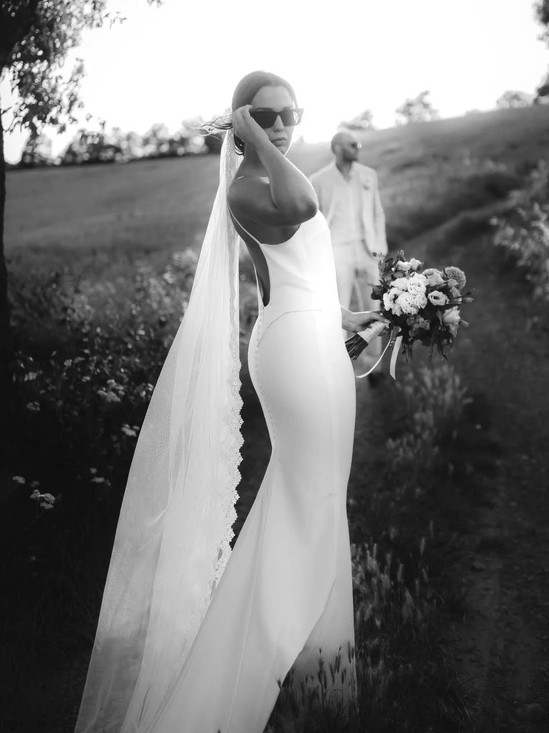 bride with sunglasses looking elegant in the countryside with the groom in the background