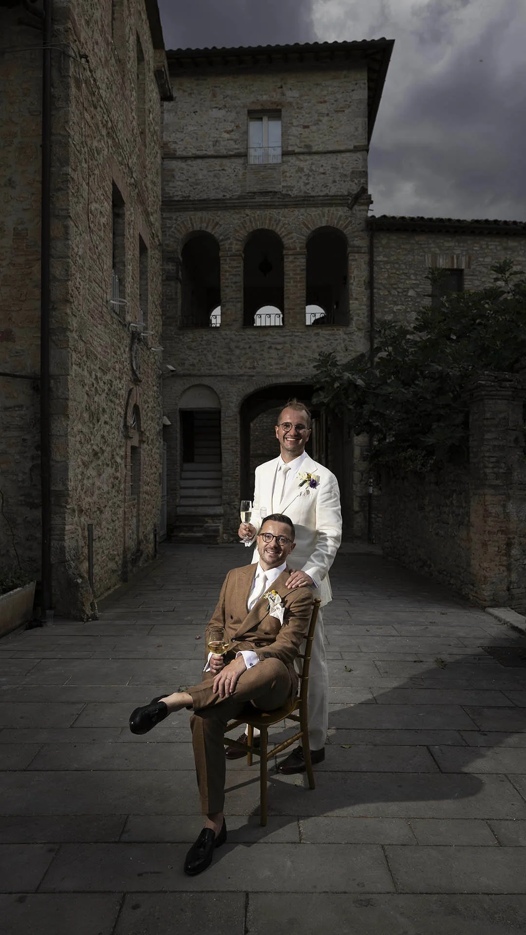 Two grooms posing with a dramatic light with glass of prosecco in the hands and a medieval building in the background