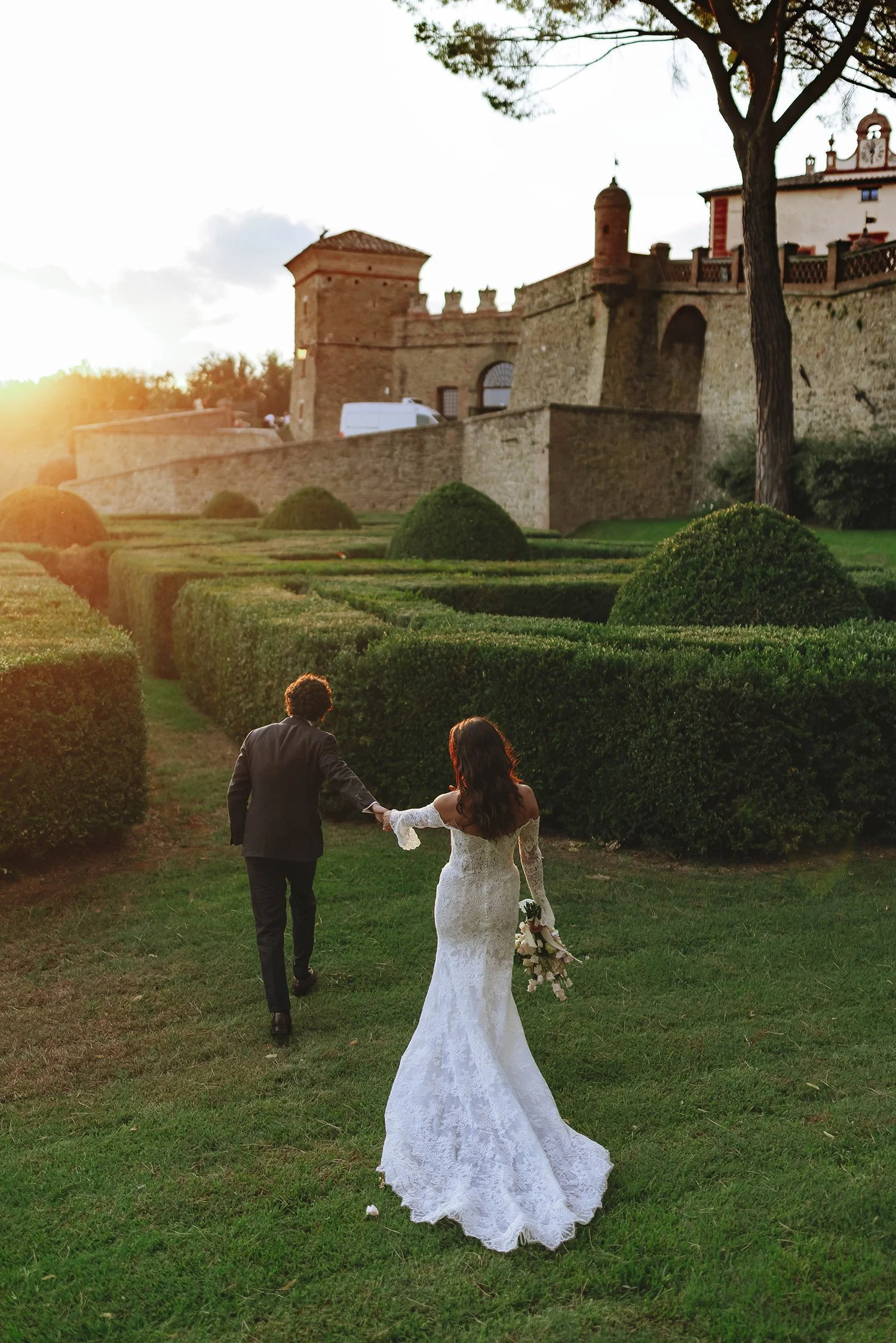 A bride and groom walking hand in hand on a grassy path near a castle, with the bride holding a bouquet of flowers, during sunset.