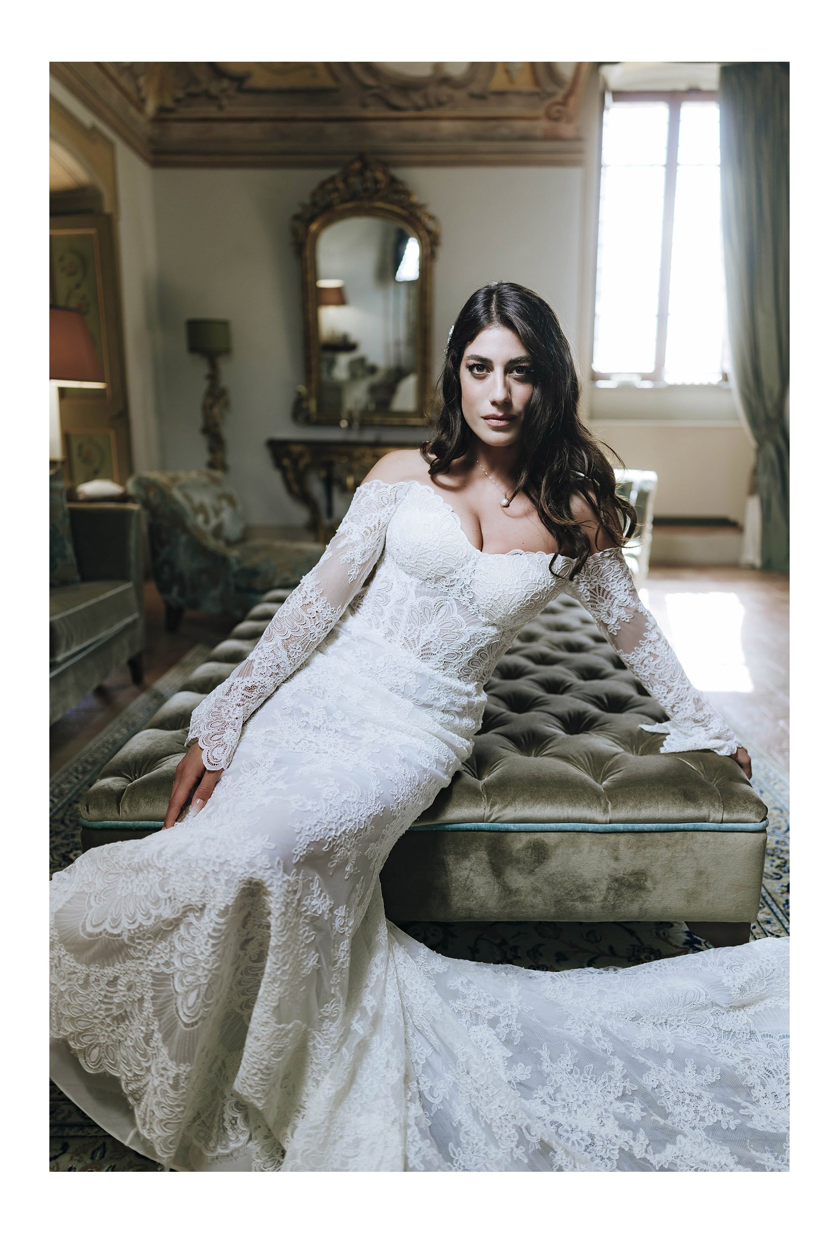 A woman in a white lace wedding dress sitting on a vintage upholstered bench in a luxurious room with classic decor, a mirror, and a window with curtains.