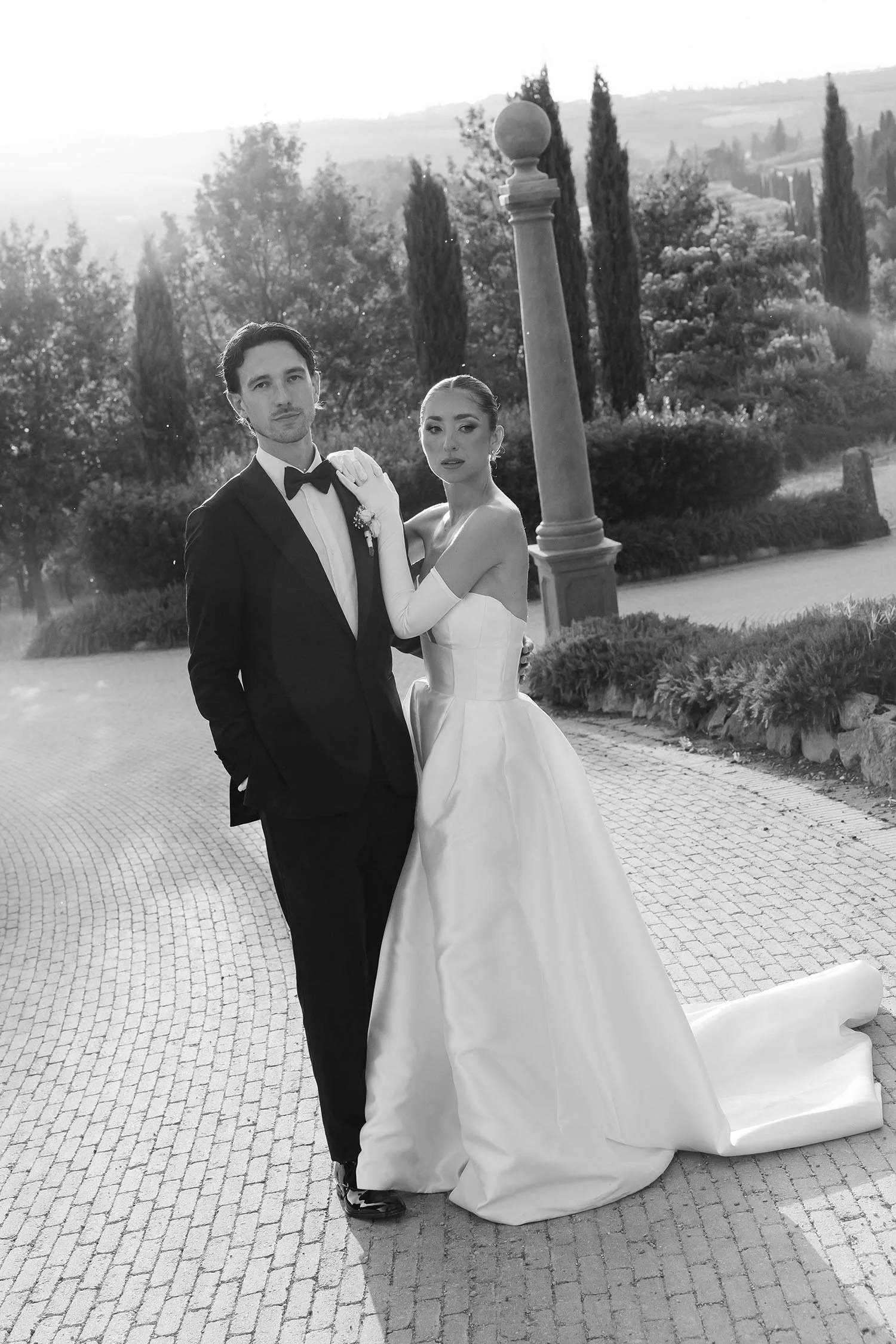 Bride and groom standing together and looking in camera in the driveway between the cypresses with the sun setting in the background