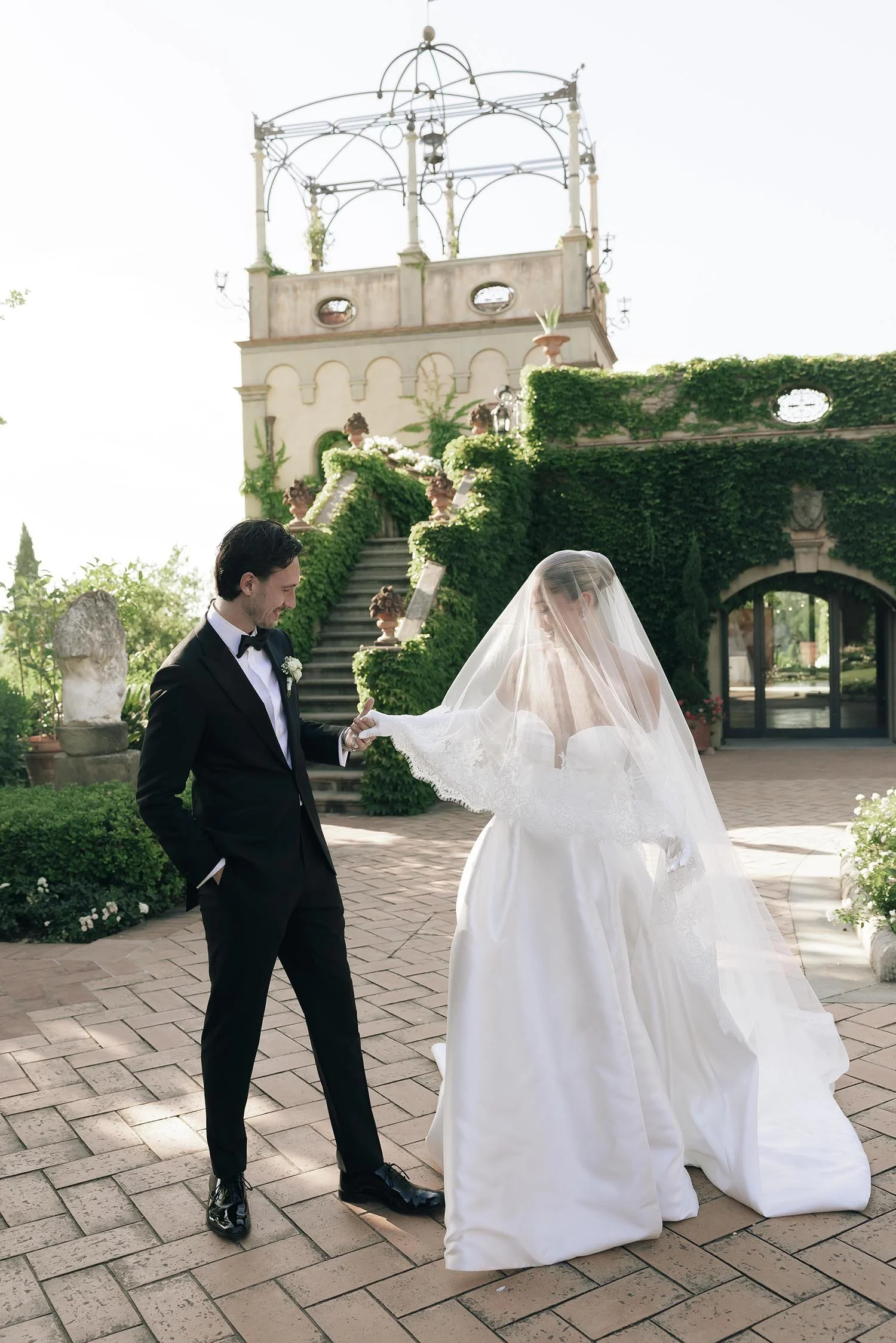 bride under the veil fixing her skirt with the groom standing beside her and the sun setting behind Tenuta Corbinaia