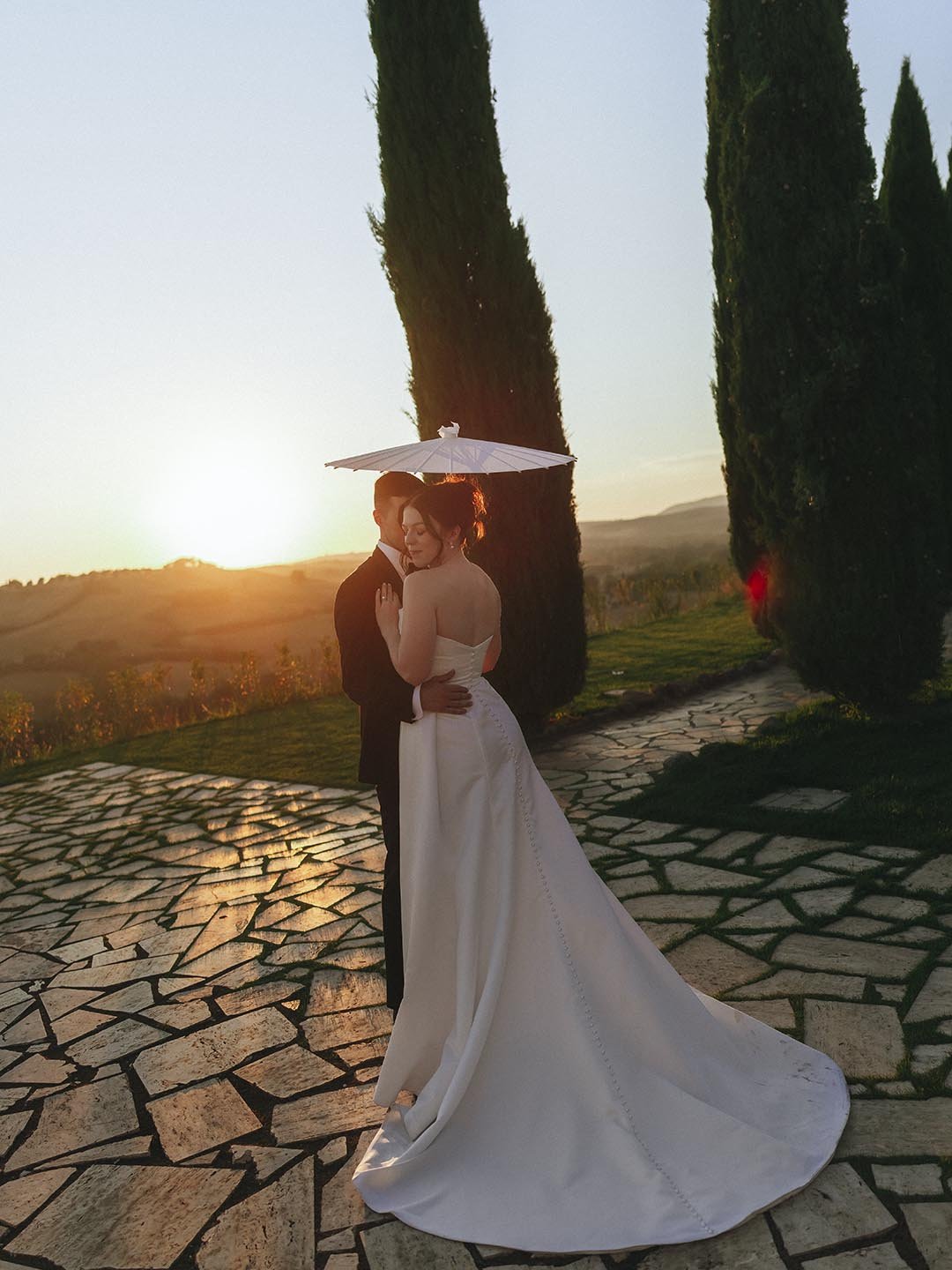 bride and groom holding each other under a parasole with the sun setting behind the cypresses at Borgo Laticastelli in Siena