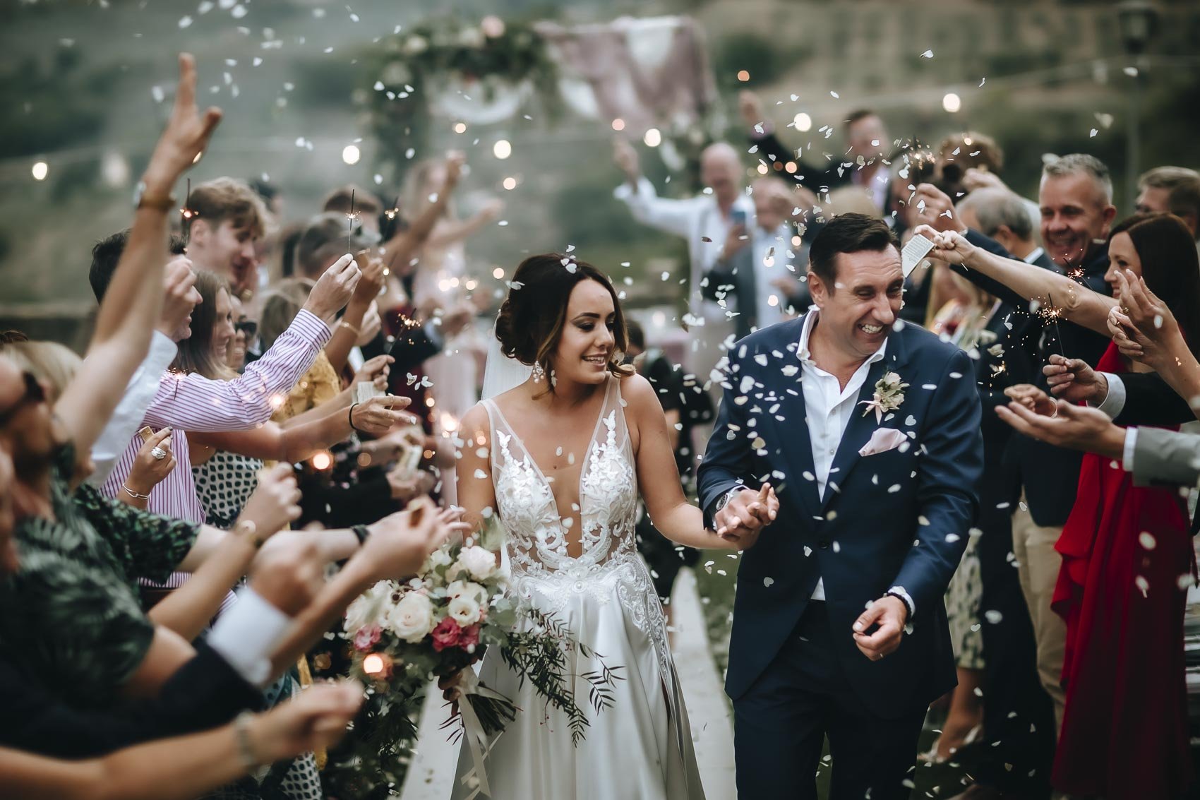 A bride and groom walk through a crowd of smiling guests holding sparklers and showering them with confetti during their wedding celebration outdoors.