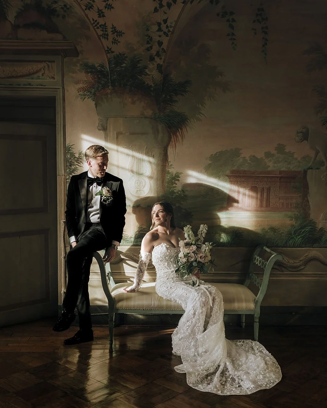 Bride sitting on the sofa with the groom beside her ina room with frescoes and hit by a dramatic light