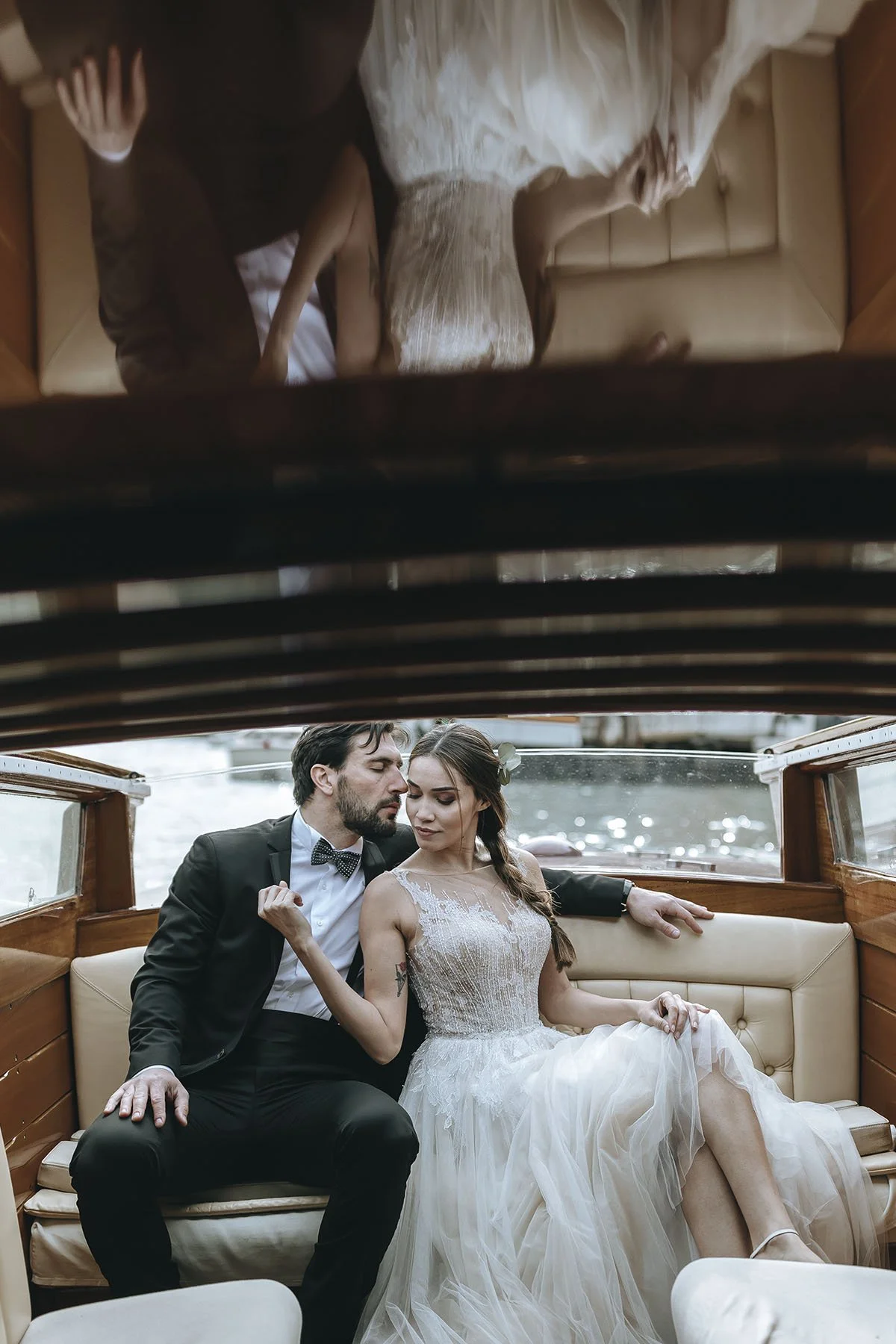 A bride and groom dressed in wedding attire sitting inside a boat, with a waterway visible through the windows behind them.