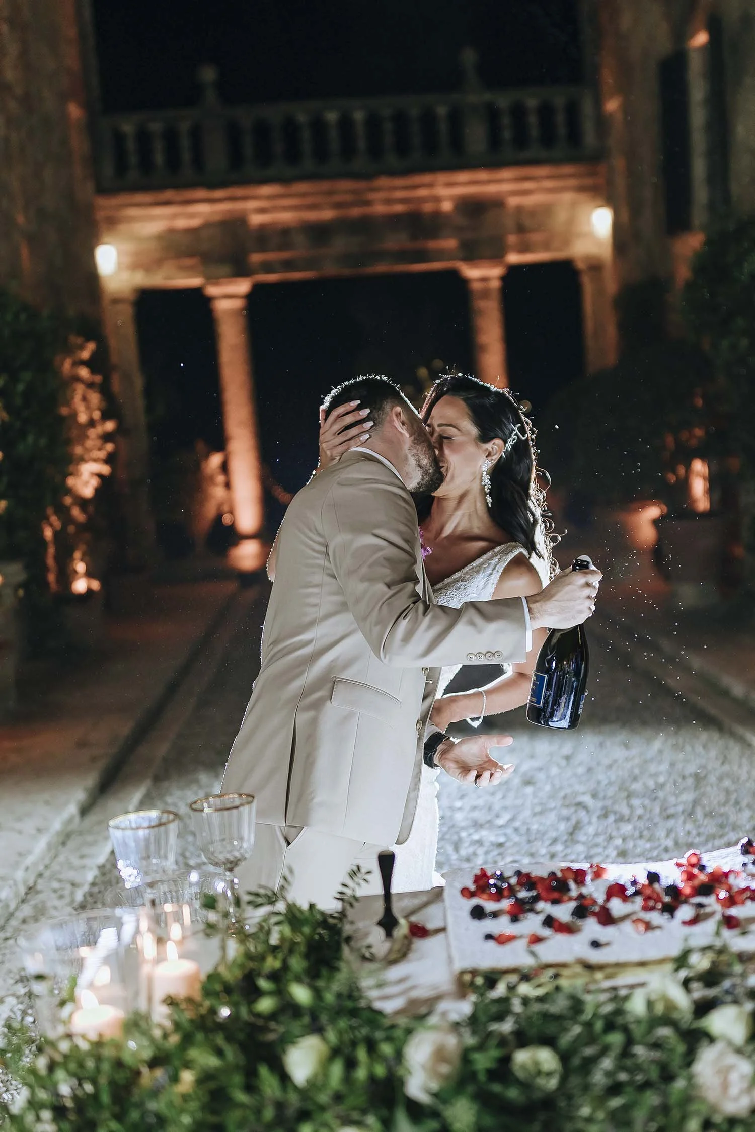 Bride and groom kiss while cutting the cake and holding a bottle of prosecco
