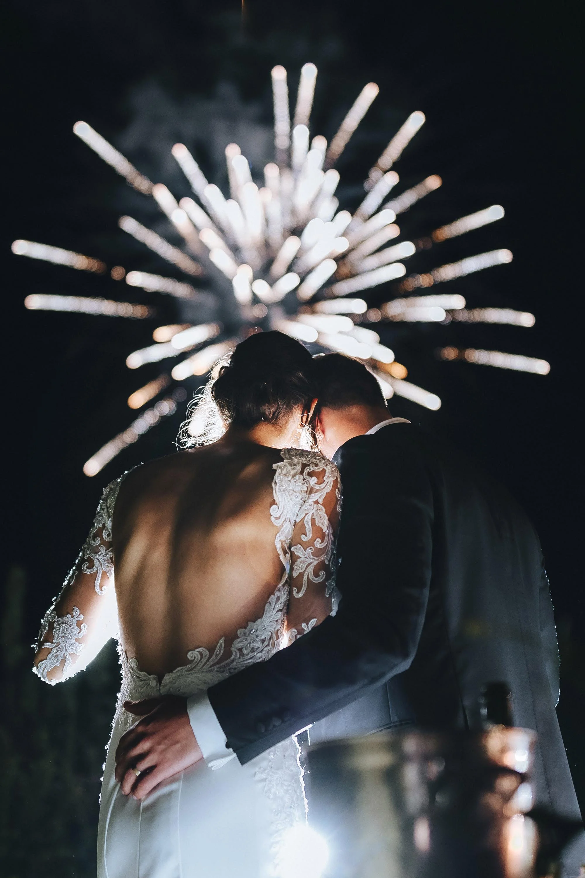 Bride and groom holding each other while cutting the cake and fireworks in the background