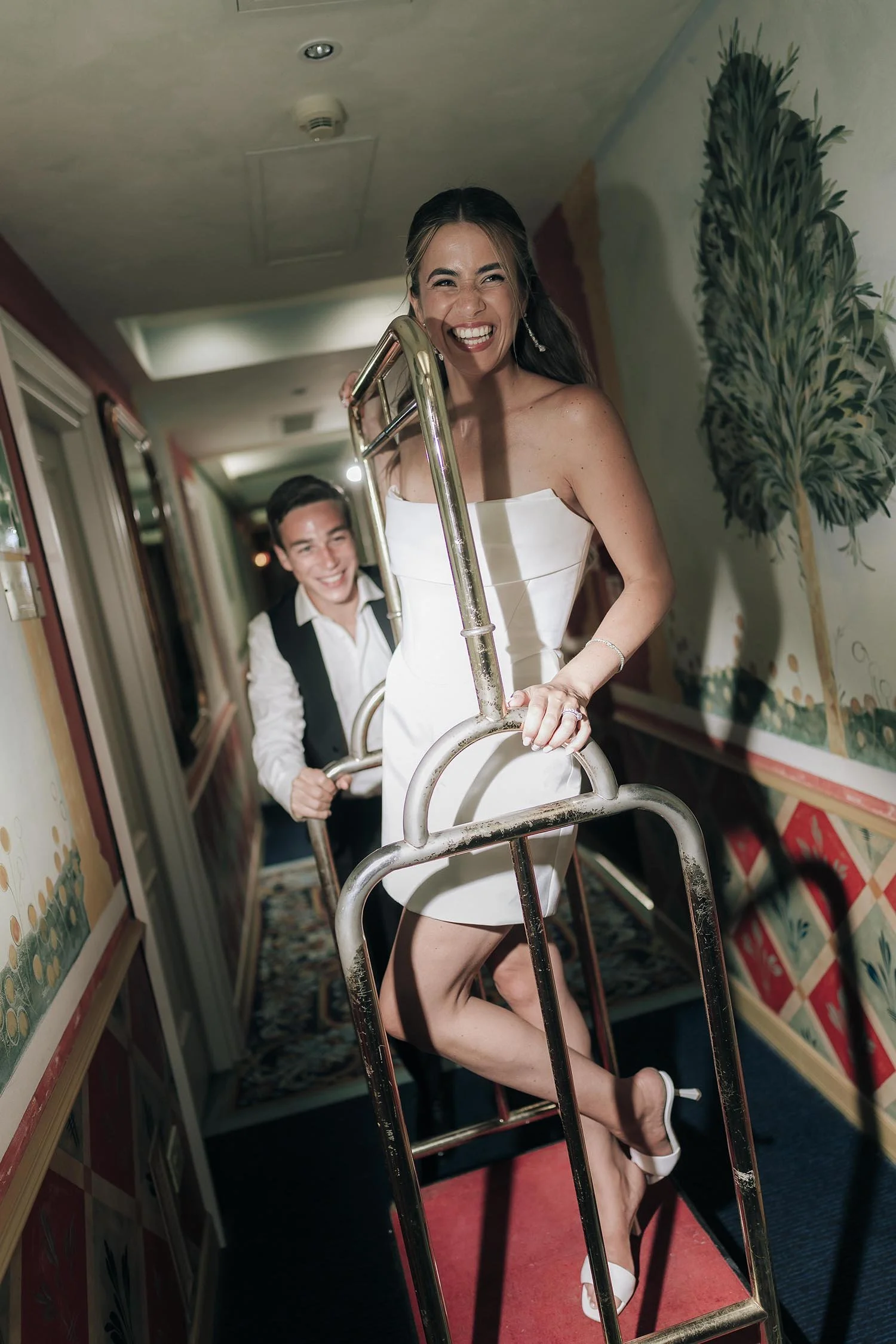 Groom carrying the bride on a bellman trolley in an hotel corridor