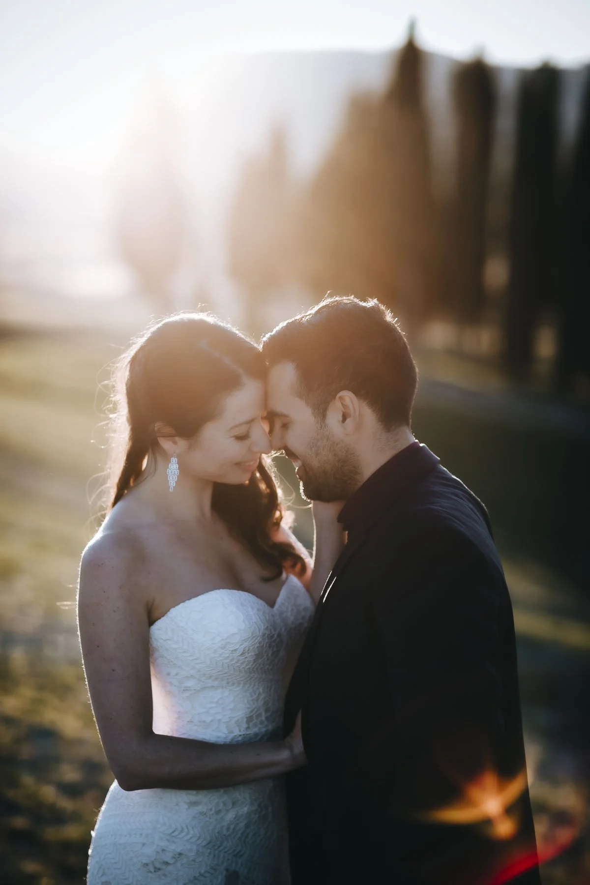 A couple is intimate with foreheads touching, standing outdoors at sunset, with the woman in a wedding dress and the man in a dark suit.