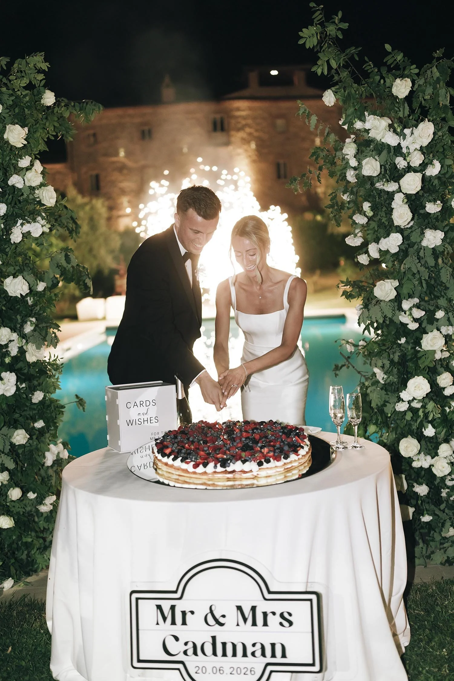 bride and groom cutting the cake by the pool with fireworks in the background