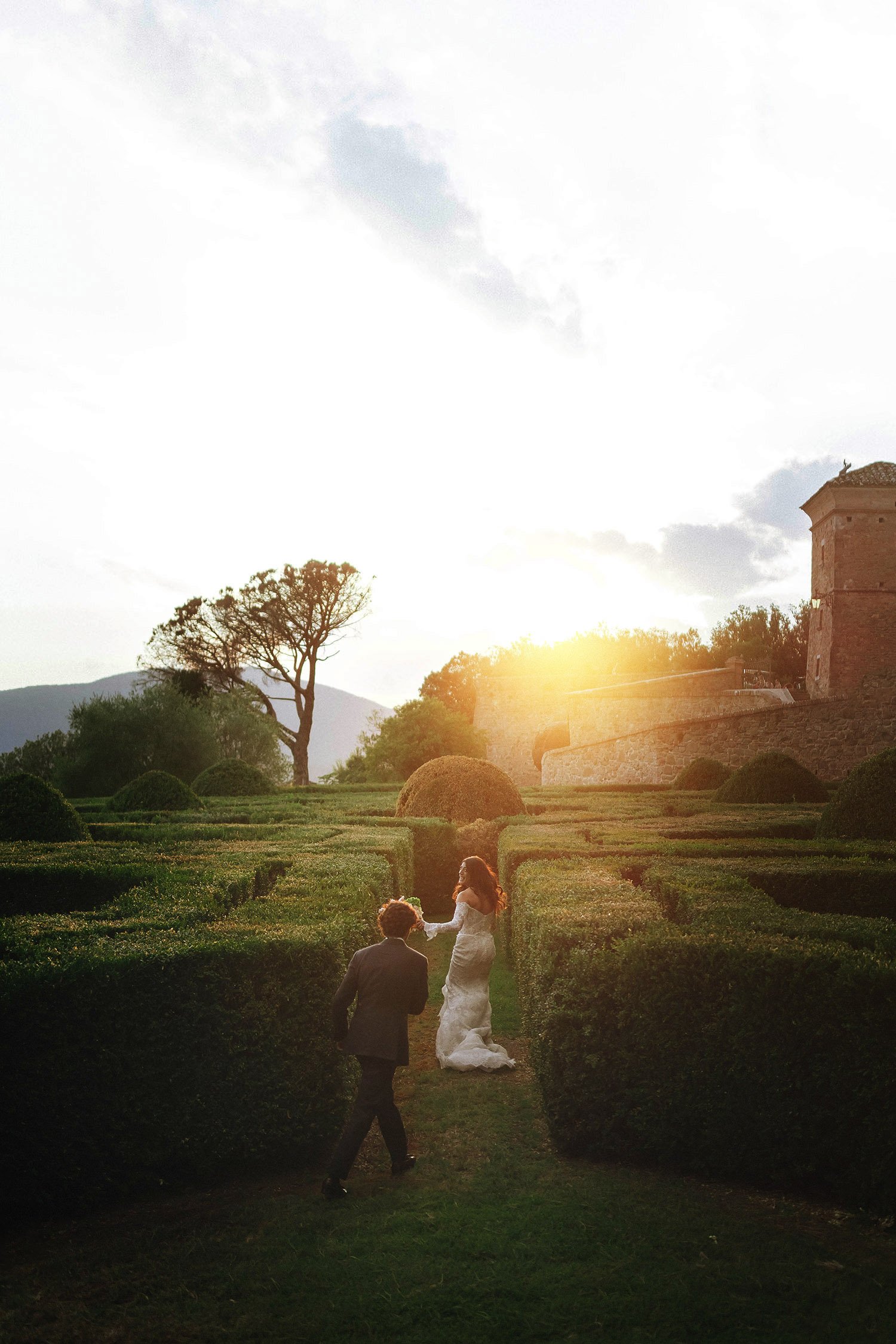 A bride and groom in wedding attire walking through a landscaped garden with neatly trimmed bushes, during sunset, with a historic building and mountains in the background.