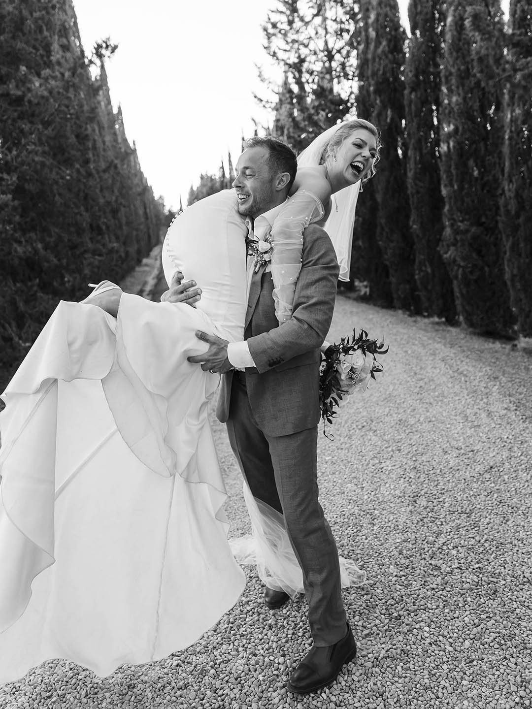 Groom holding the bride on his shoulder along the cypresses