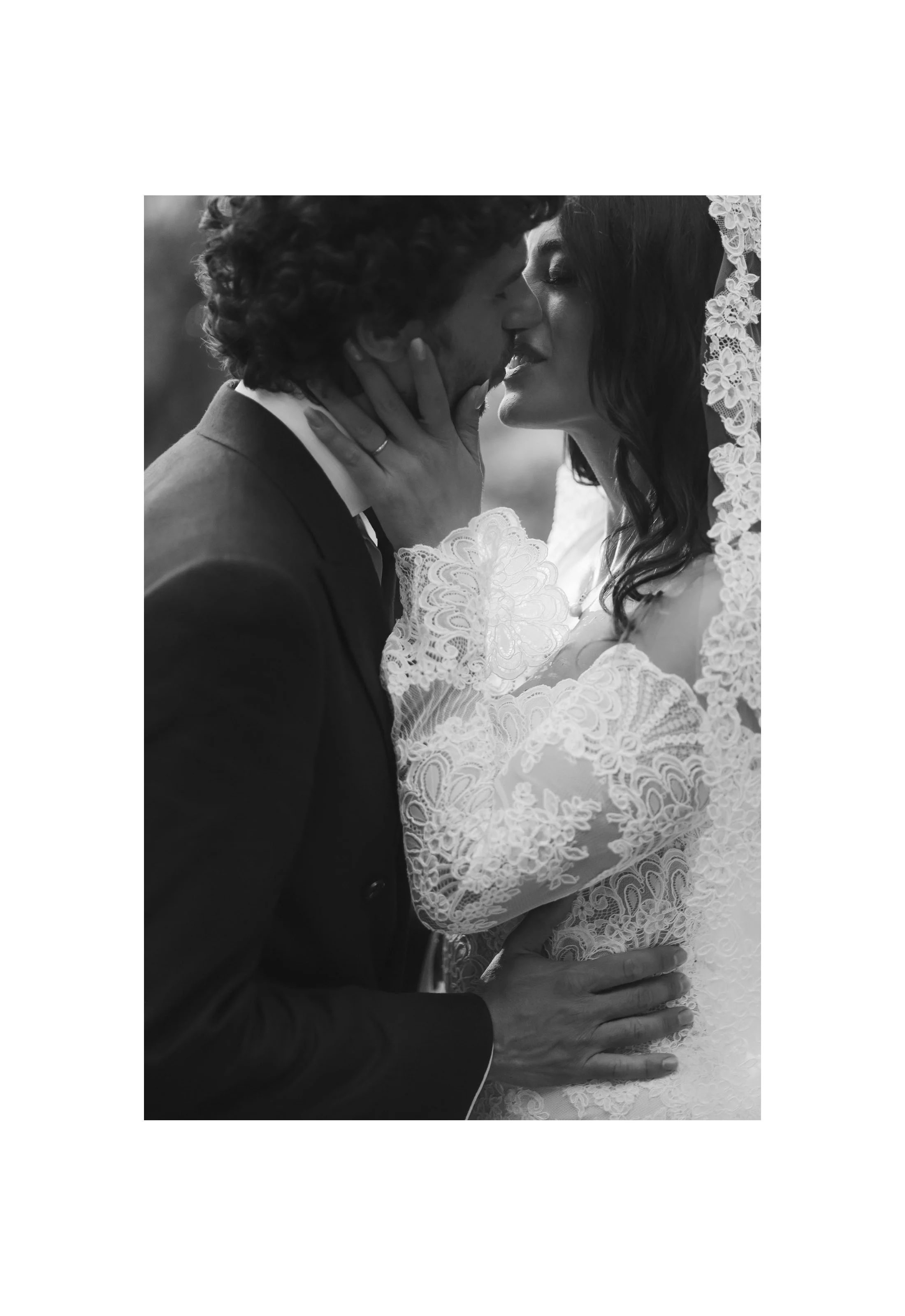 A black and white photo of a couple kissing, with the woman wearing lace wedding attire and the man in a suit.