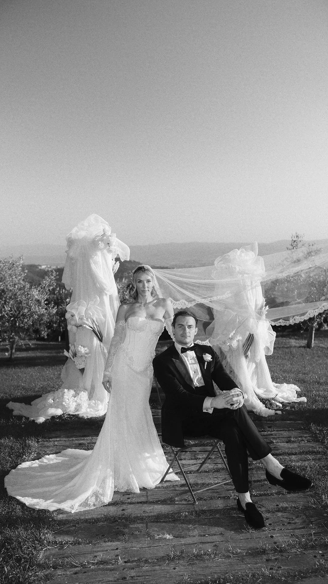 editorial portrait of bride and groom posing in the garden of Castello di Petrata, the groom is sitting on a chair and the bride stands beside him with the veil flowing in the wind
