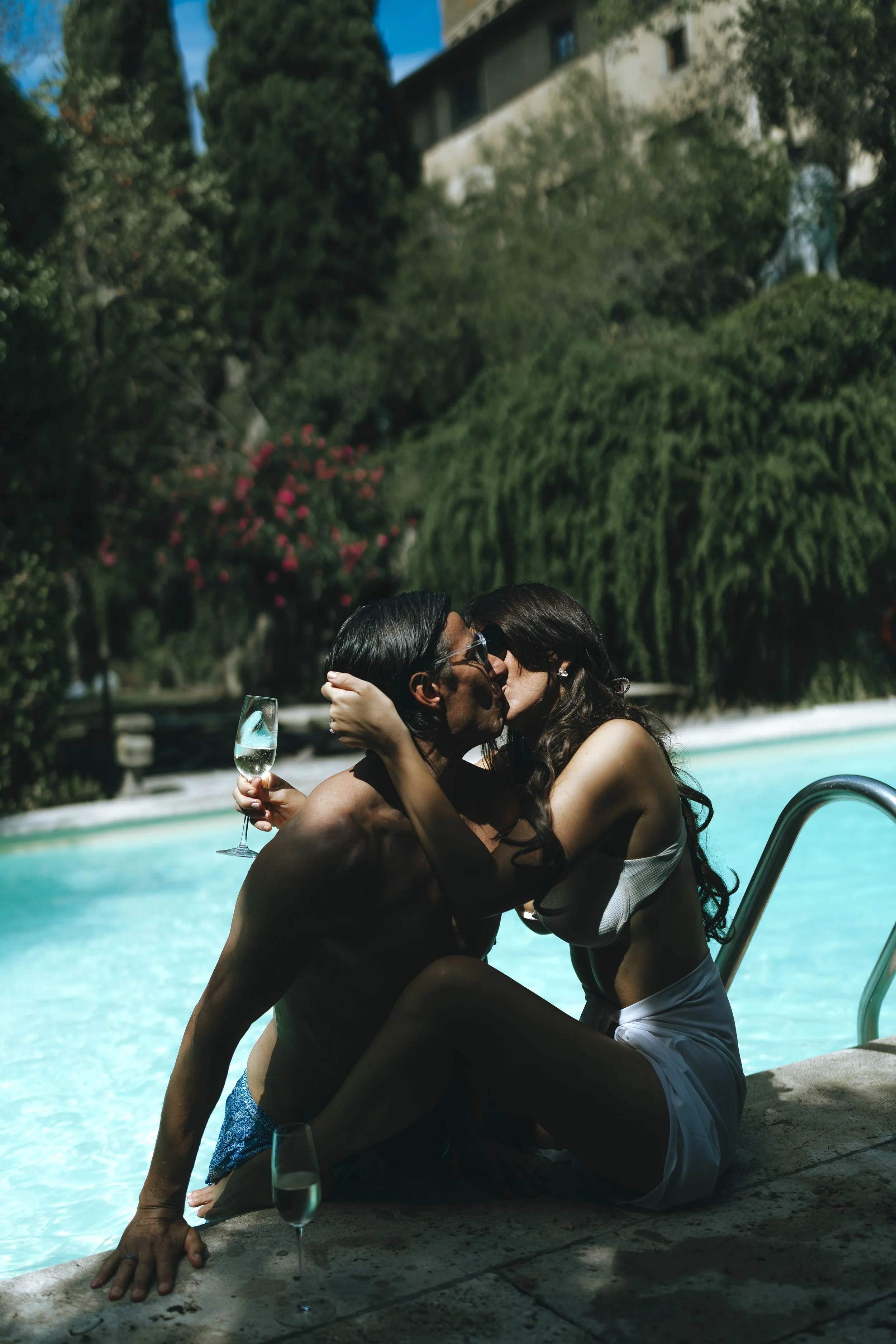 Bride and groom kiss each other in the swimming pool holding glass of prosecco