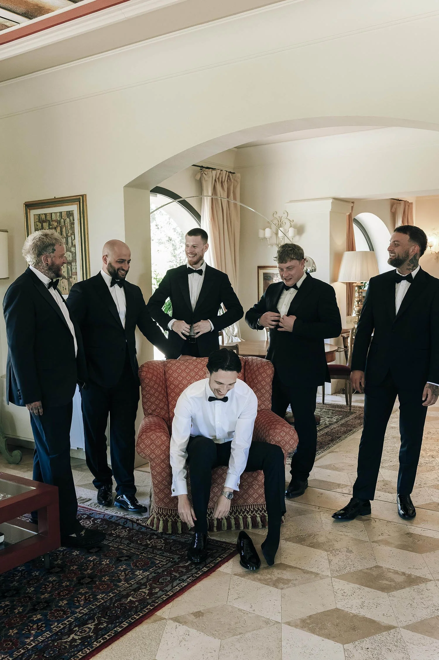 the groom sitting on the armchair fixing his shoes with all the groomsmen standing around him in the living room at Tenuta Corbinaia