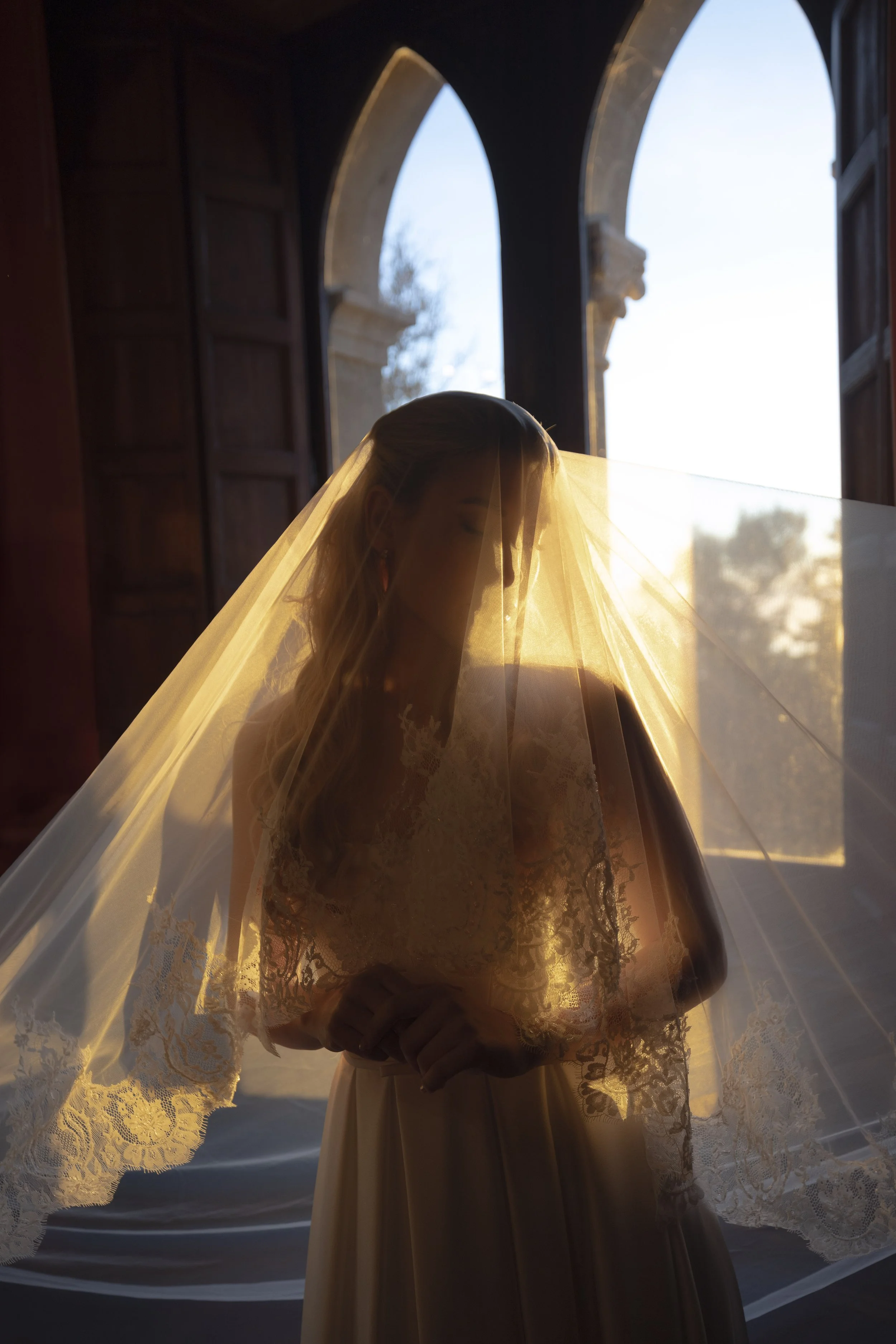 A bride standing indoors near a large window with sunlight illuminating her silhouette, wearing a lace wedding dress and veil. Photo by Rellini art studio documentary wedding photographers in Italy