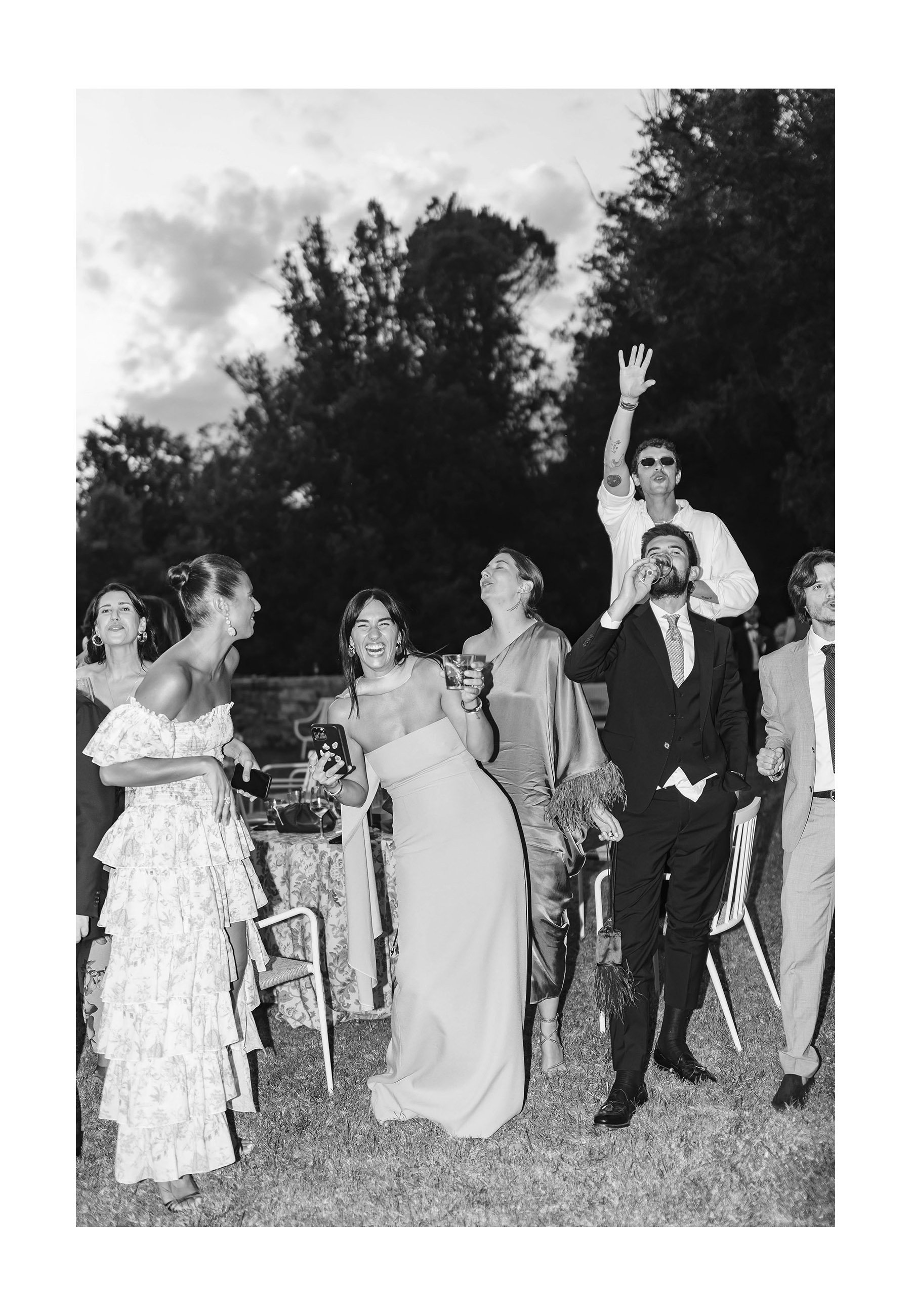 People at an outdoor nighttime celebration, some holding drinks, smiling, and raising their hands, with trees and cloudy sky in the background.