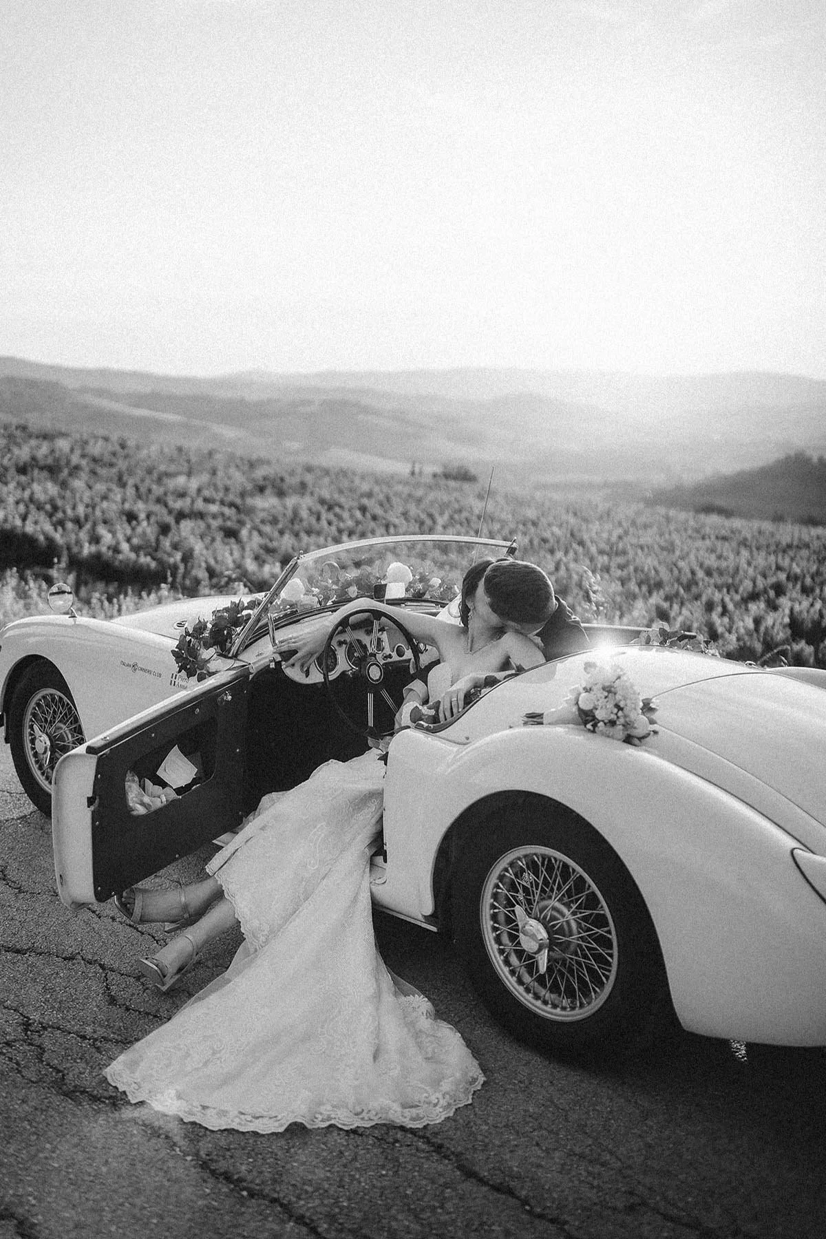 bride and groom kiss each other in a vintage convertible white car with the sun setting on the vineyards near Todi in Umbria