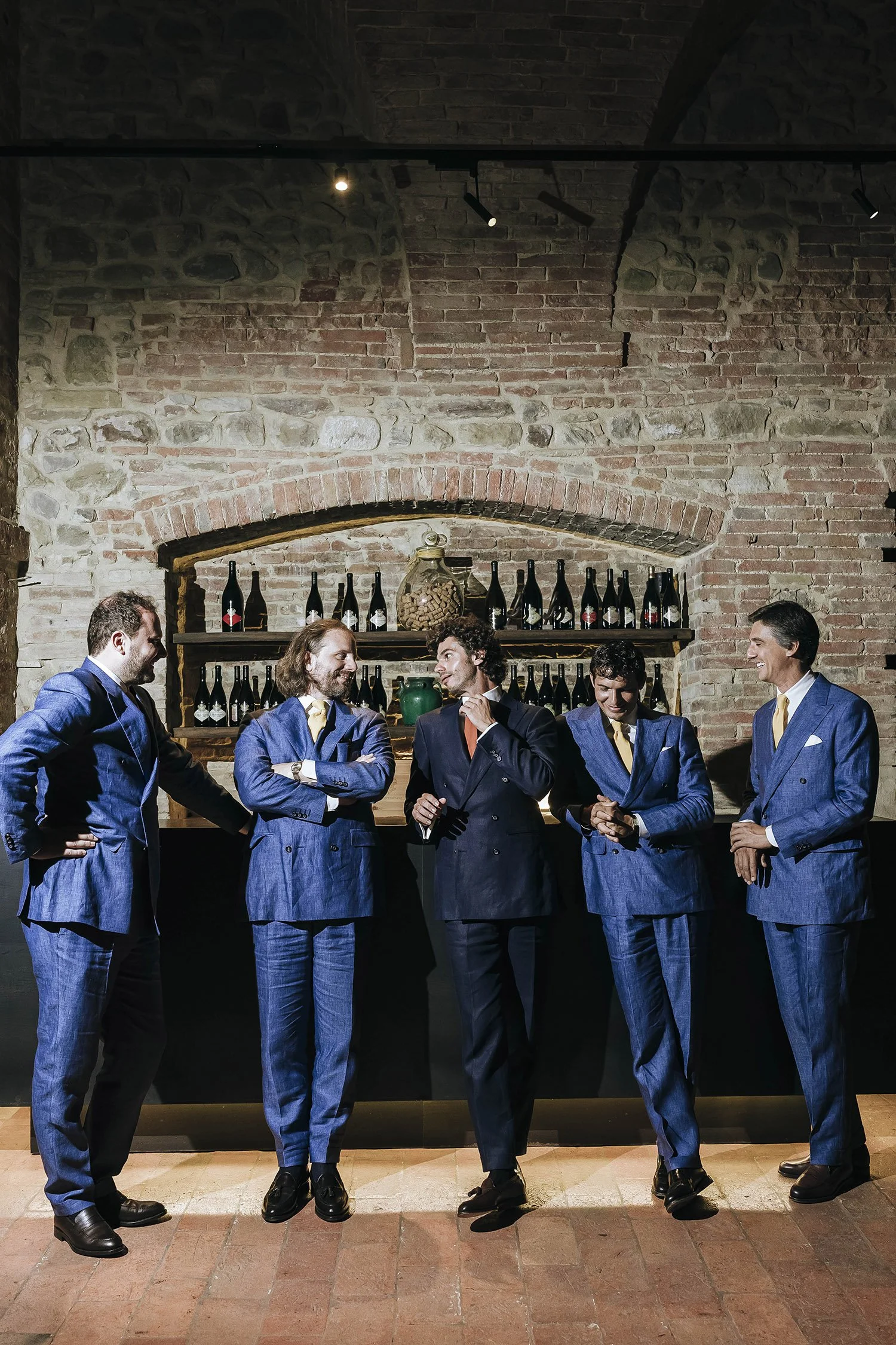 Five men in blue suits are standing together, talking and smiling in front of a bar with one holding a glass, against a brick wall background with shelves of bottles.