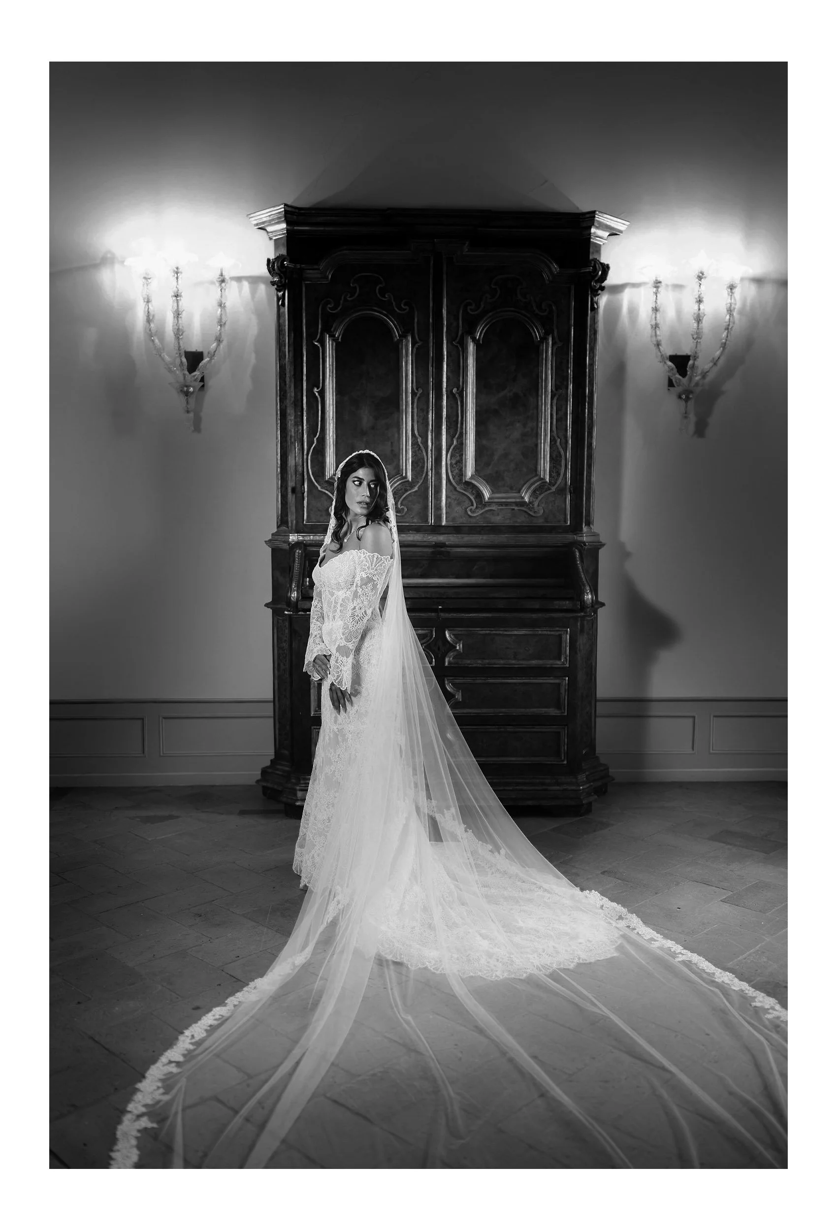 A woman in a wedding dress with a long train stands in a dimly lit room with ornate wall sconces and an antique wooden cabinet in the background, captured in black and white.
