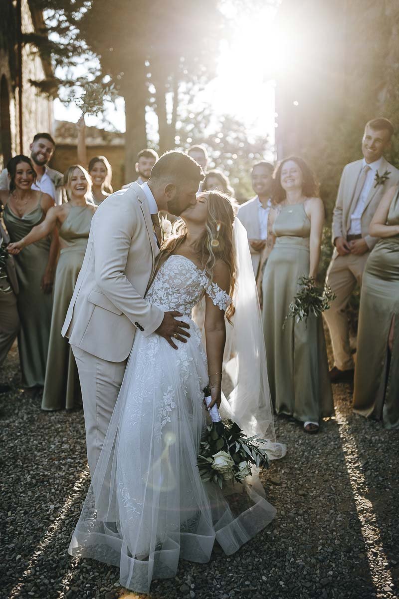 Destination wedding at Ristonchi castle near Florence in Tuscany.  bride and groom kiss each other with their bridal party in the background hit by a warm sunset light