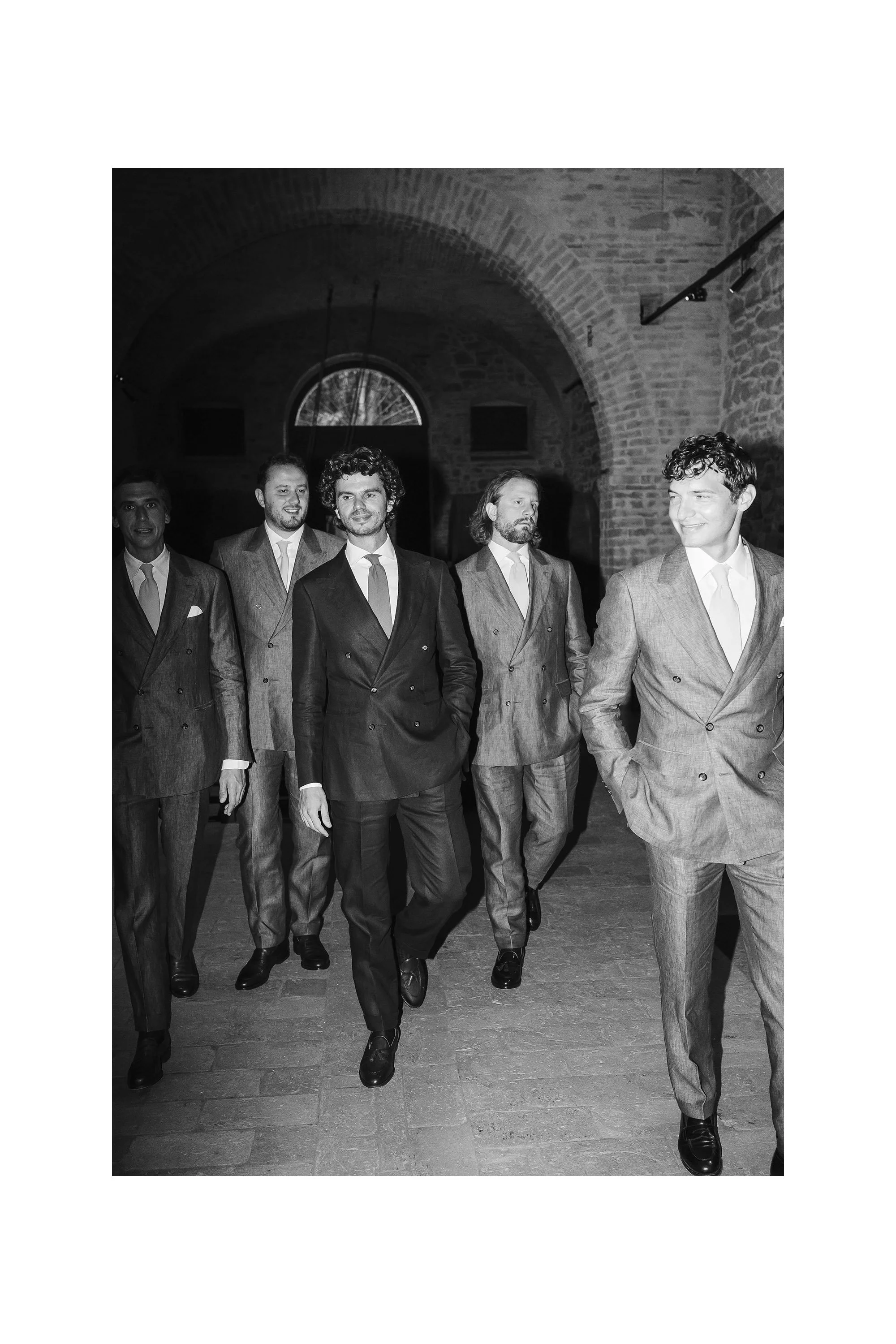 Group of five men in suits walking in a brick archway with a window in the background.