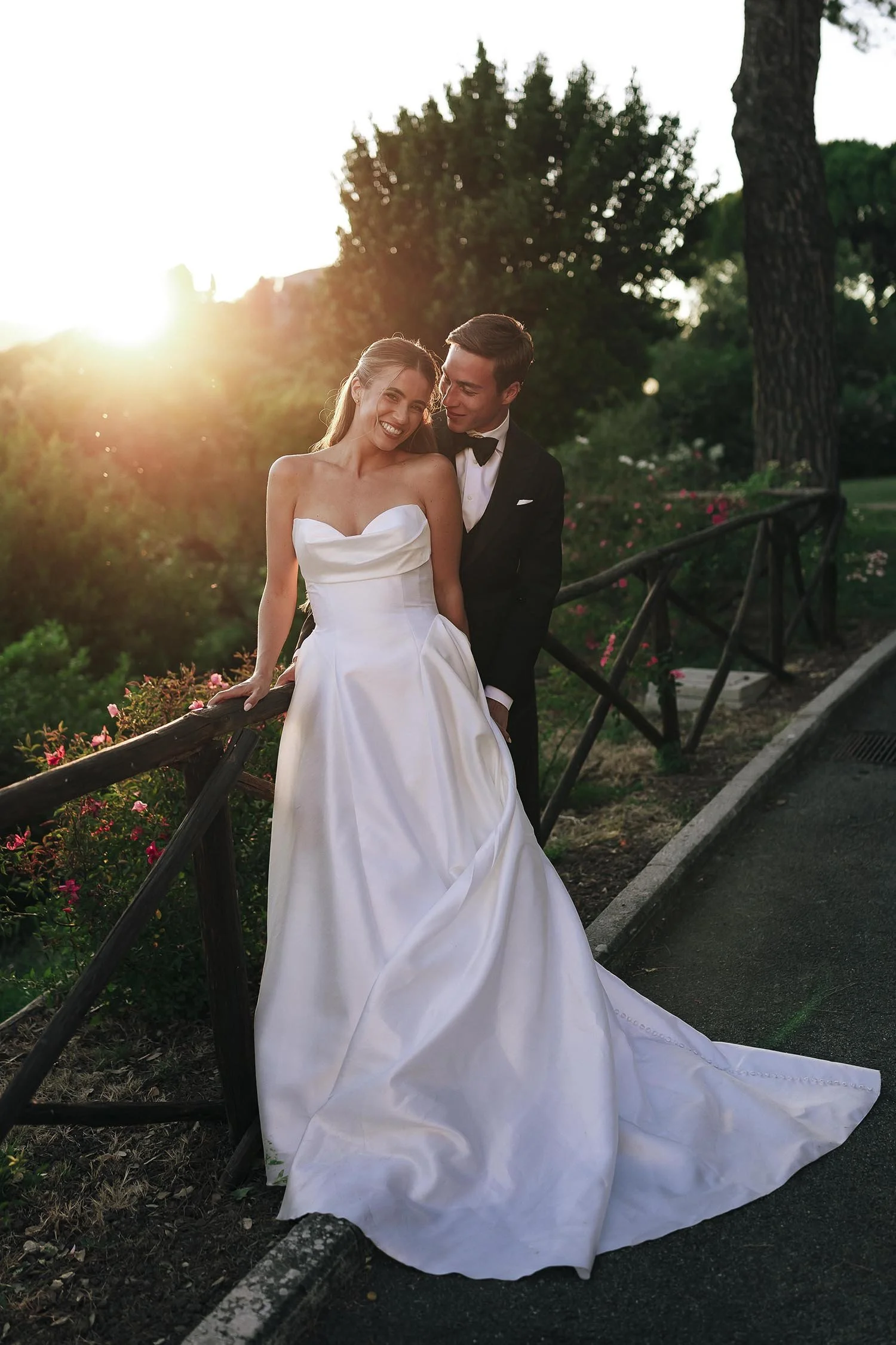 groom holding the bride by a fence with the golden light of the sun setting in the background