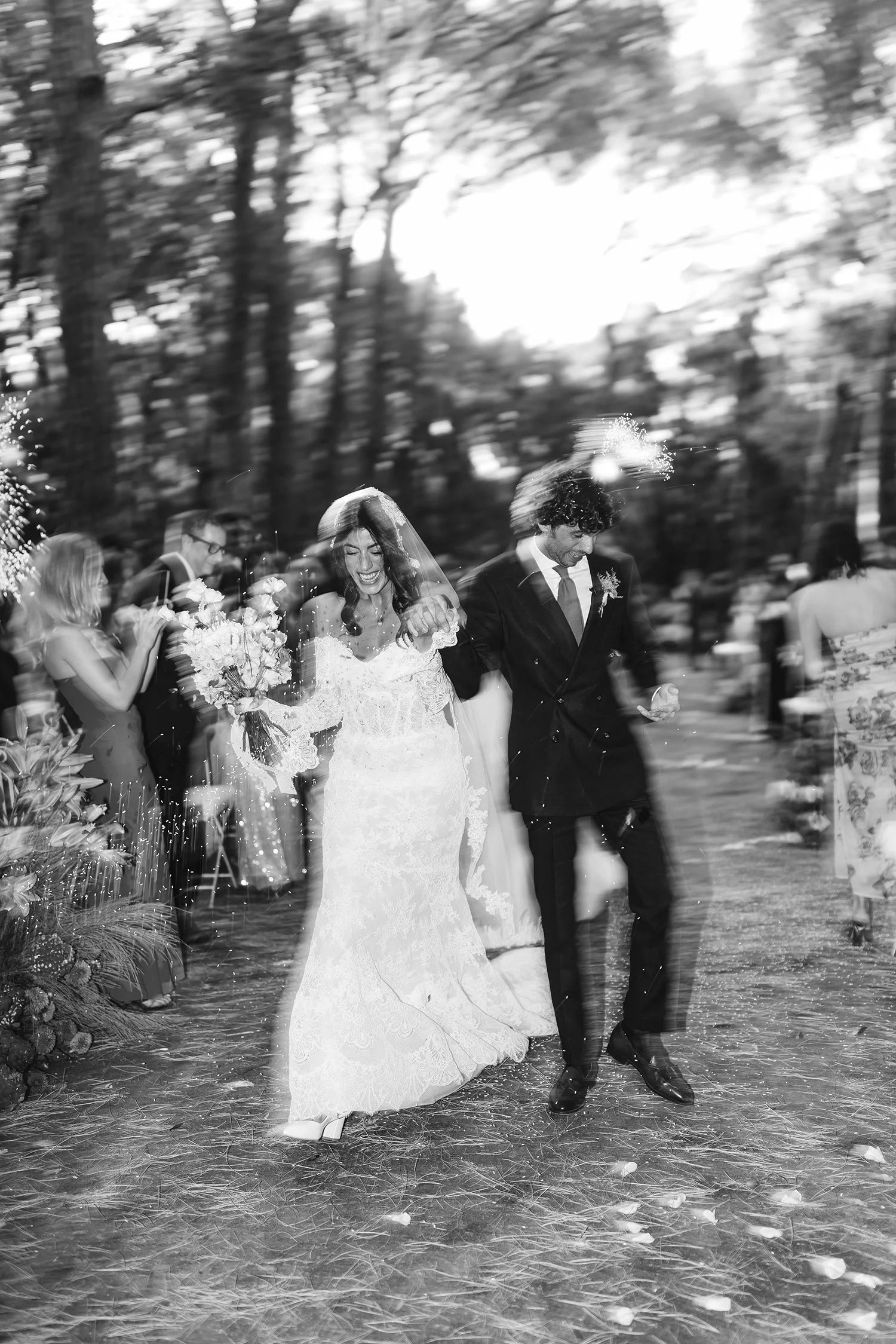 Black and white photo of a newlywed couple walking down the aisle, smiling, with wedding guests around them, outdoor setting with trees.