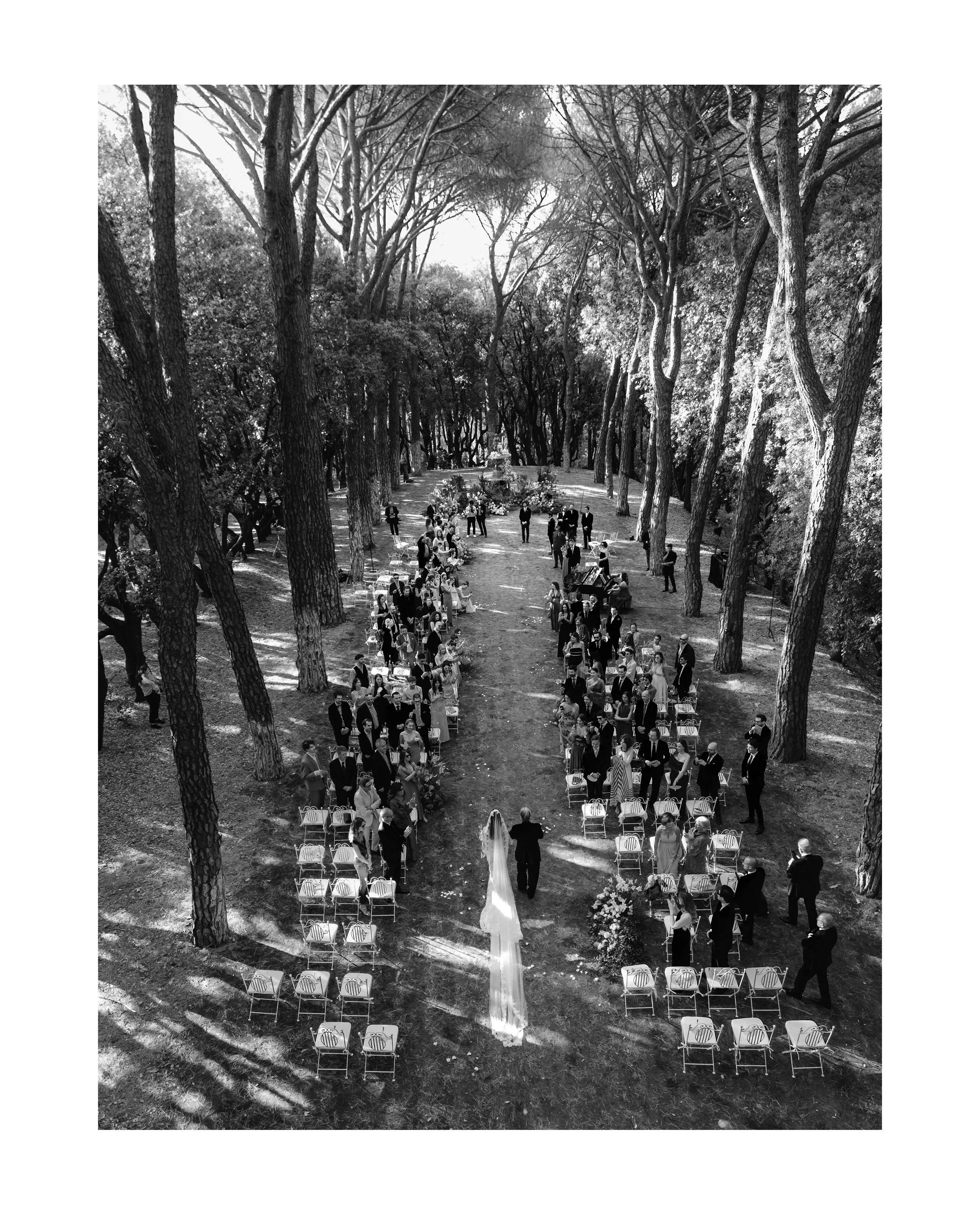 A black-and-white photograph of an outdoor wedding ceremony set in a wooded area with tall trees. The bride in a white dress and veil stands at the altar with her partner, while guests are gathered around, seated and standing along the aisle.