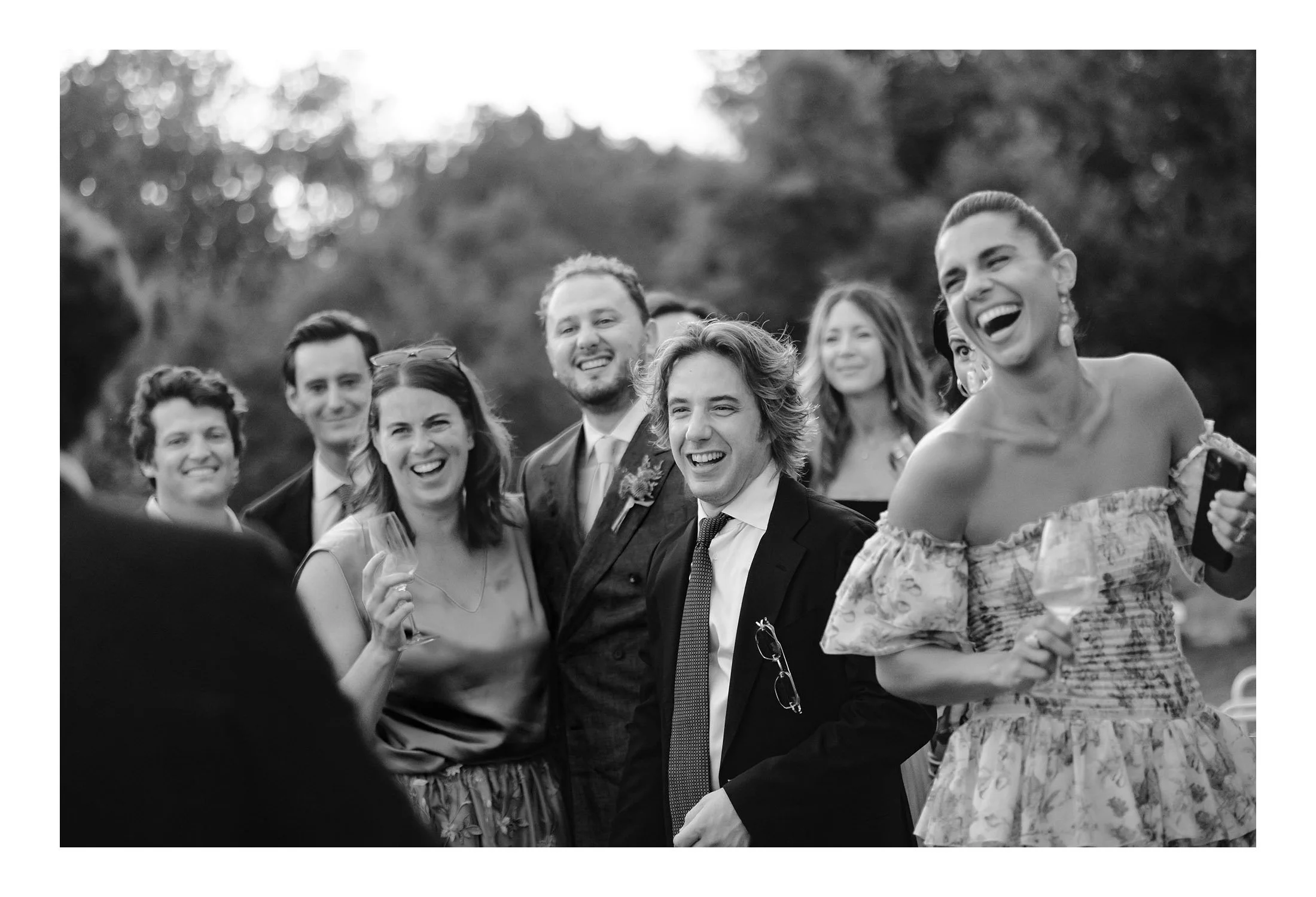 Black and white photo of a group of people at an outdoor event, smiling and laughing, with trees in the background.