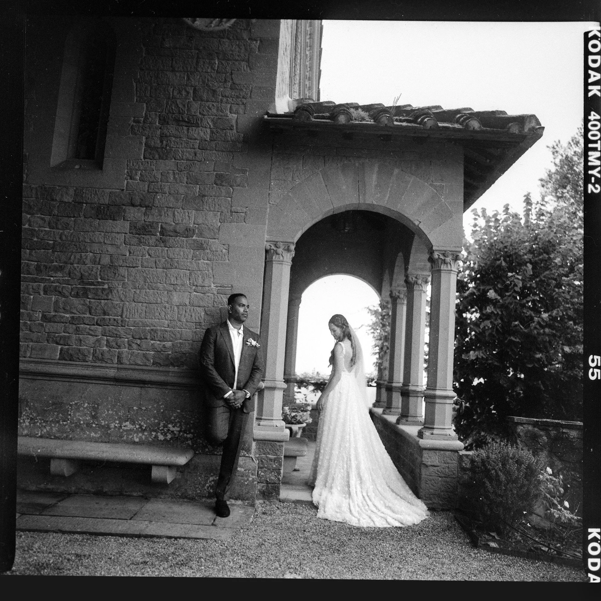 A black and white photo of a bride and groom outdoors under a stone archway. The groom is standing, dressed in a dark suit, with his hands clasped in front. The bride is in a long wedding gown, standing slightly to the right, with her head tilted dow