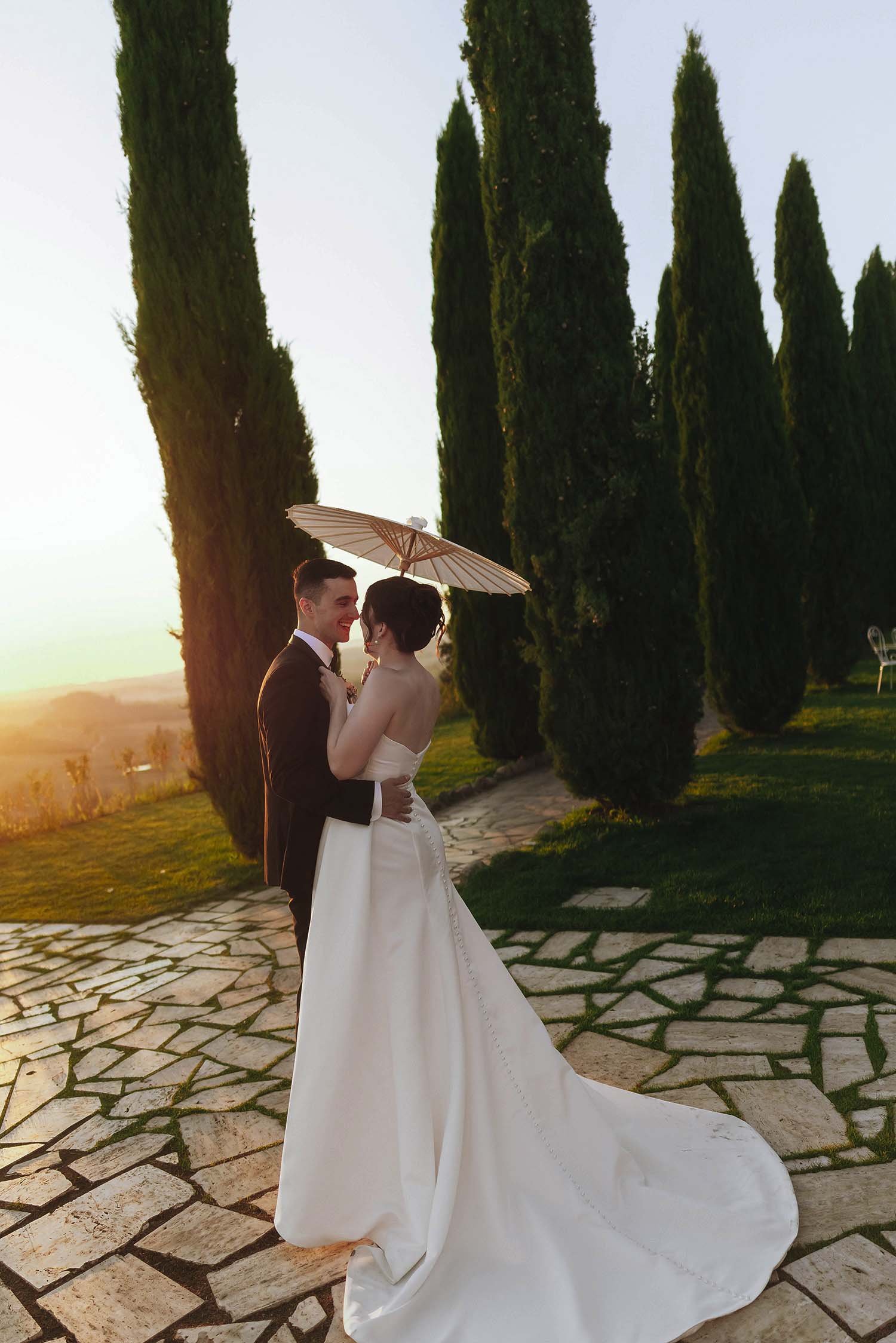 Bride and groom holding each other holding a parasole with the sun setting behind the cypresses